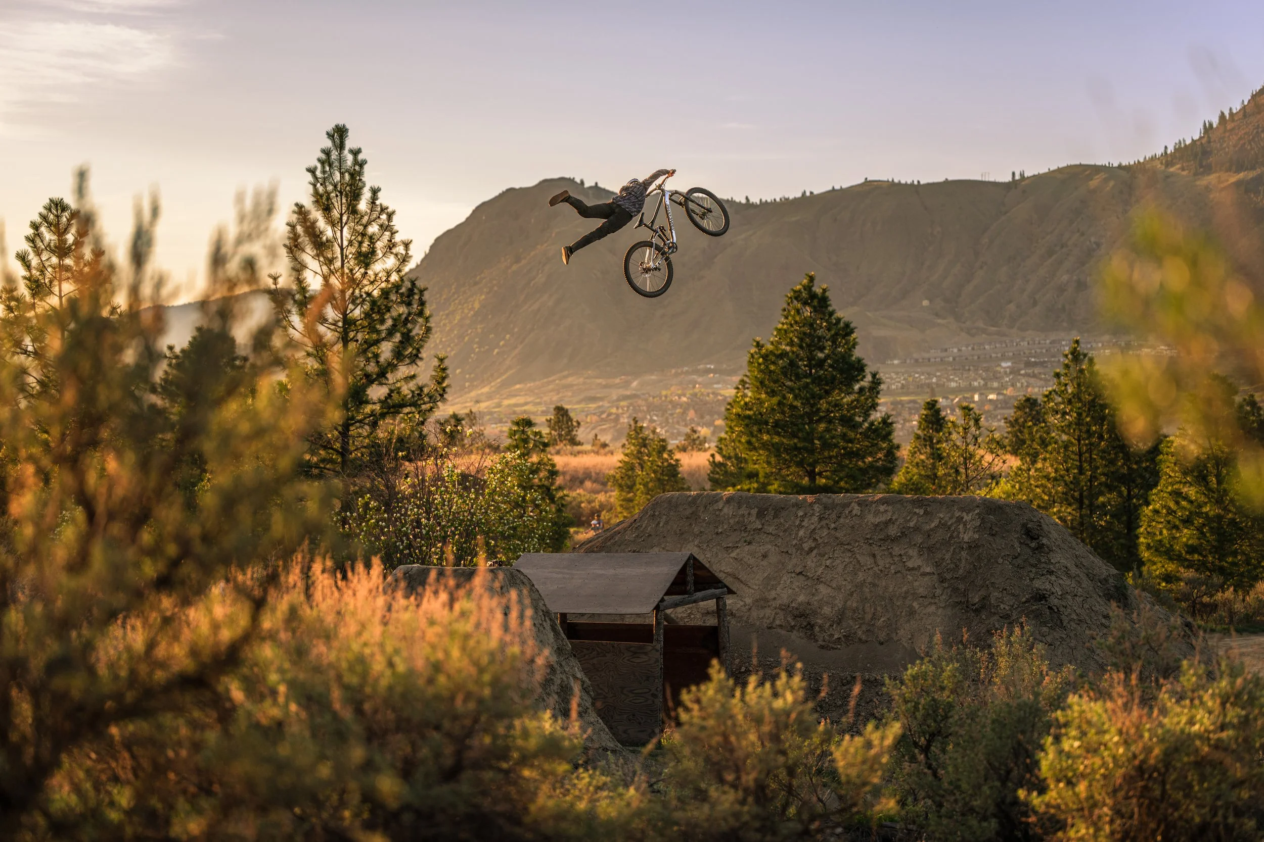 Person performing a trick on a mountain bike in a dirt jump park during sunset with mountains and trees in the background.