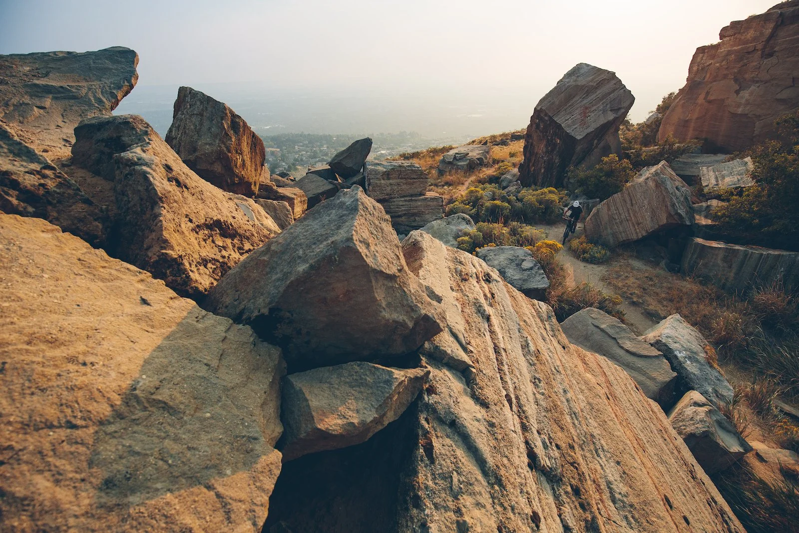 A mountain trail scattered with large rocks and boulders, with a cyclist riding along the narrow path, surrounded by dry grass and bushes, with a hazy cityscape in the distance at sunset.