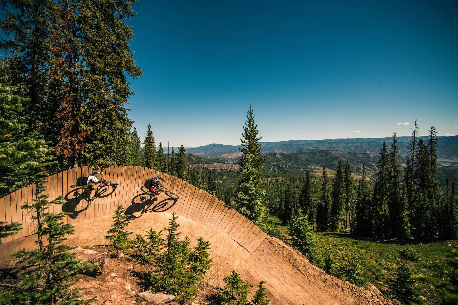Two mountain bikers riding on a curved dirt track in a forested mountainous landscape with evergreen trees and distant mountain ranges under a clear blue sky.