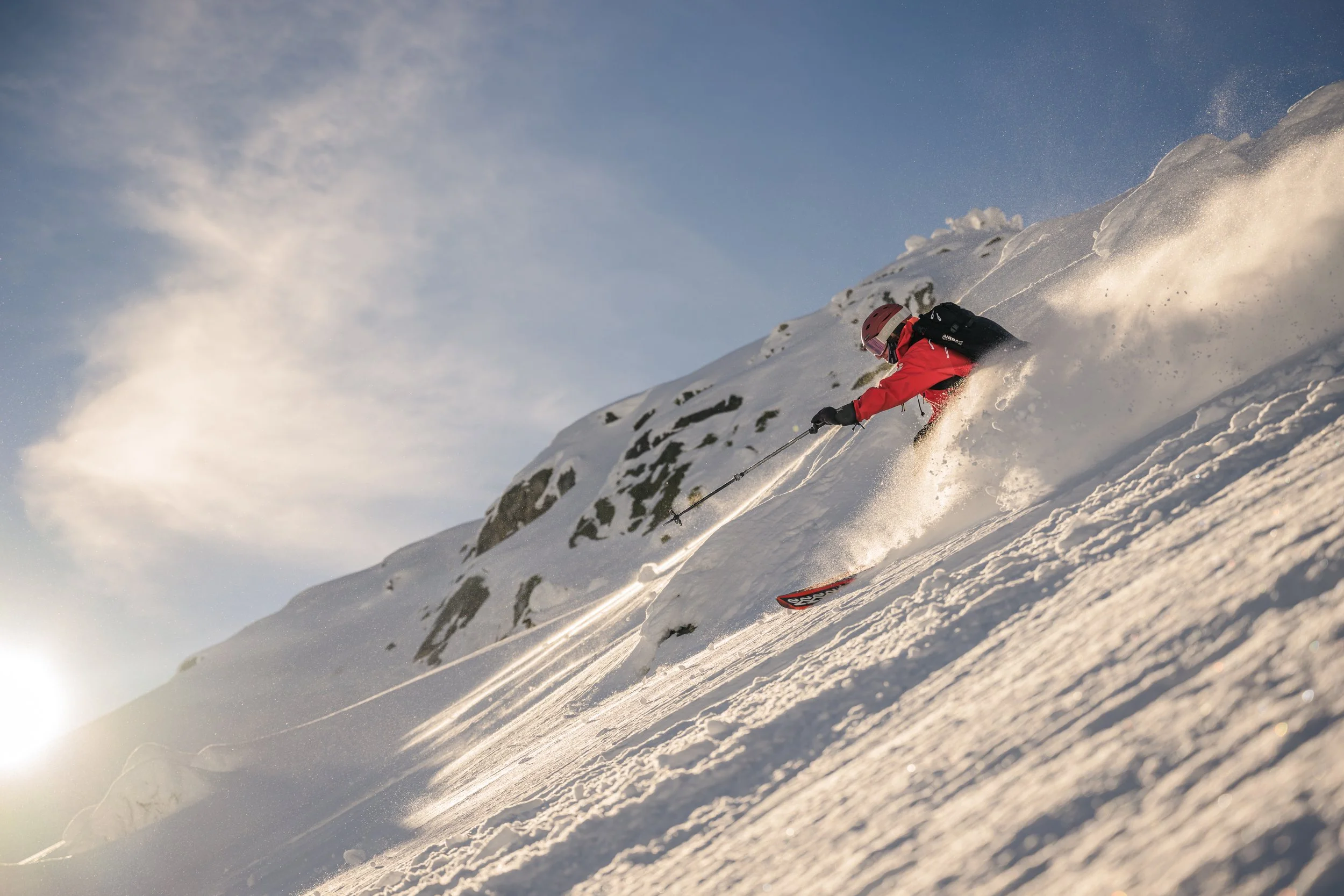 Skier in red jacket and helmet skiing down snowy mountain slope, kicking up snow with a backdrop of partly cloudy sky.