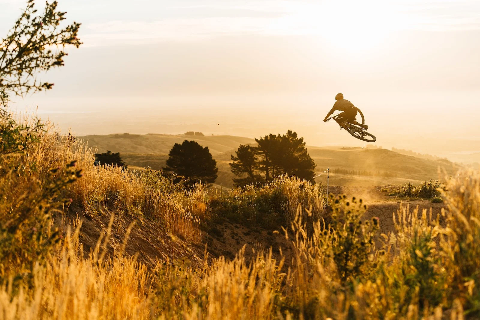 A person riding a mountain bike and airborne over a dirt trail during sunset, surrounded by grassy hills and trees.