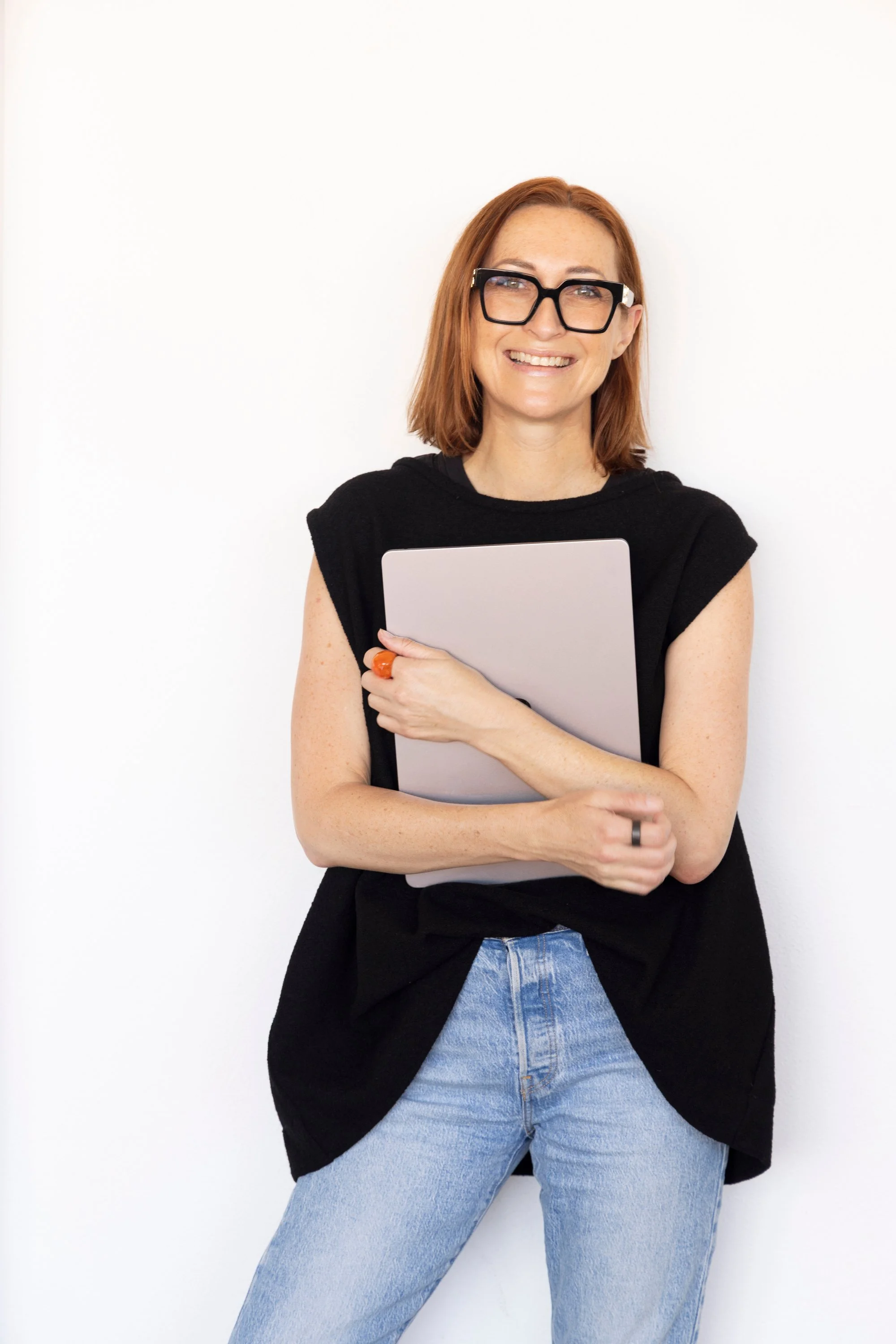 A woman with red hair, glasses, and a white shirt sitting at a desk with an open gray MacBook, a smartphone, and a pink case, smiling at the camera.