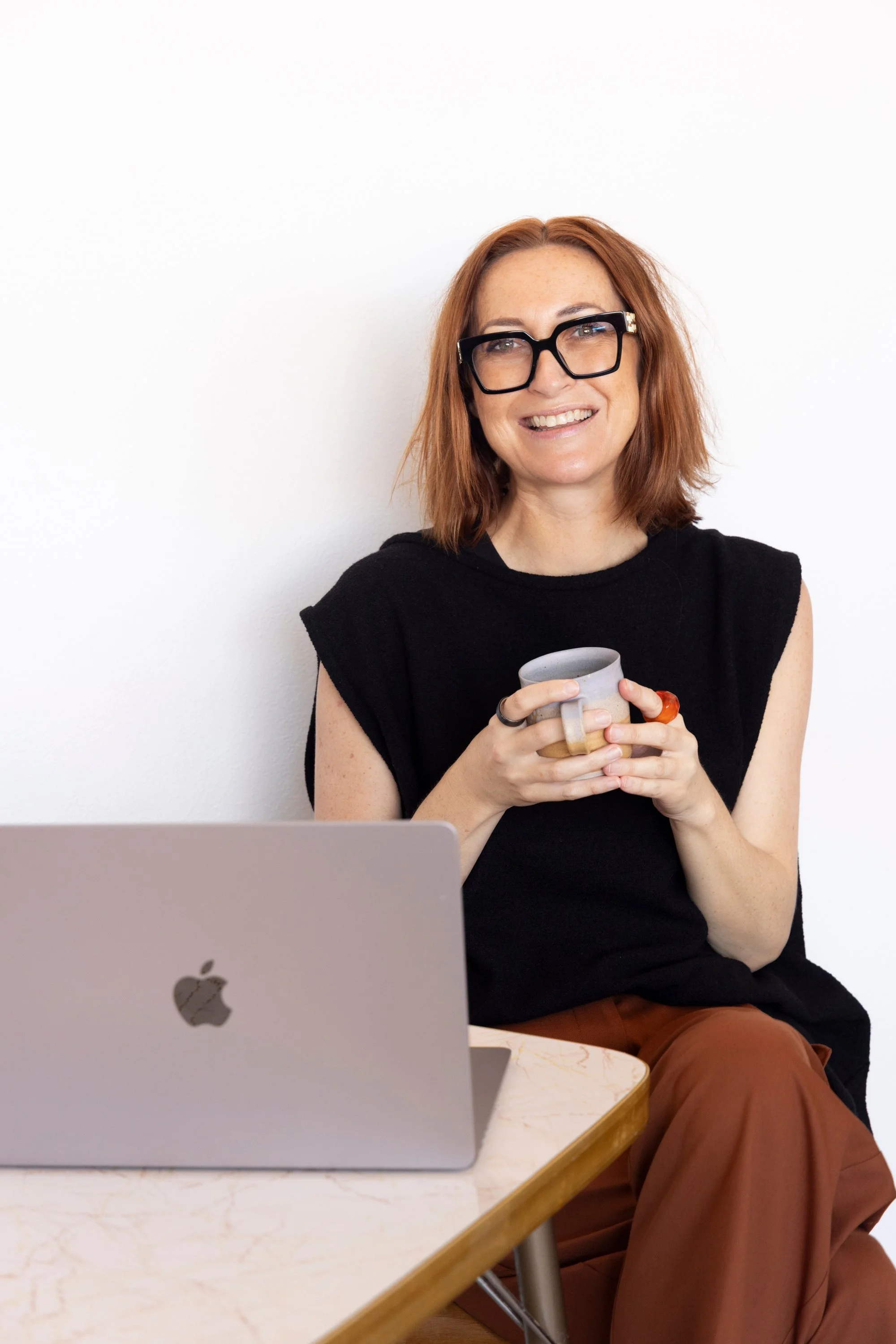 A woman with shoulder-length red hair, wearing black glasses and a sleeveless black top, sitting at a table and holding a coffee mug, with a laptop on the table in front of her, smiling against a plain white wall.