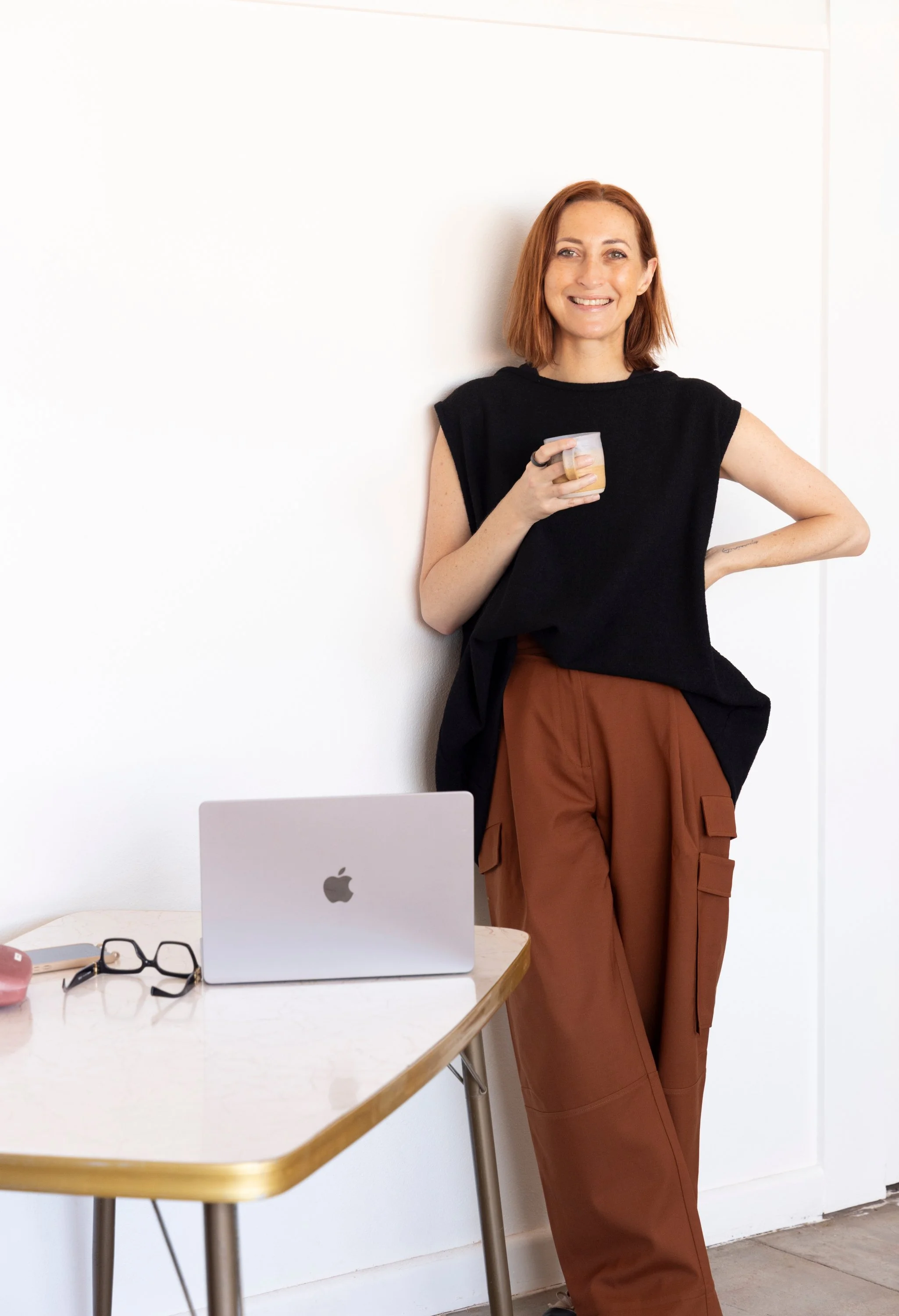 A woman with red hair, wearing glasses, a black sleeveless top, and light blue jeans, holding a laptop and smiling.