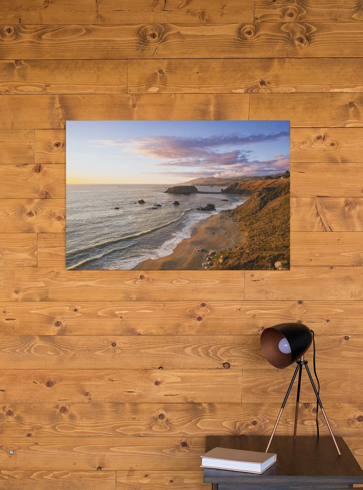 A framed landscape photograph of a coastal scene hanging on a wooden wall, with a black desk lamp and a closed book on a dark wooden surface below.