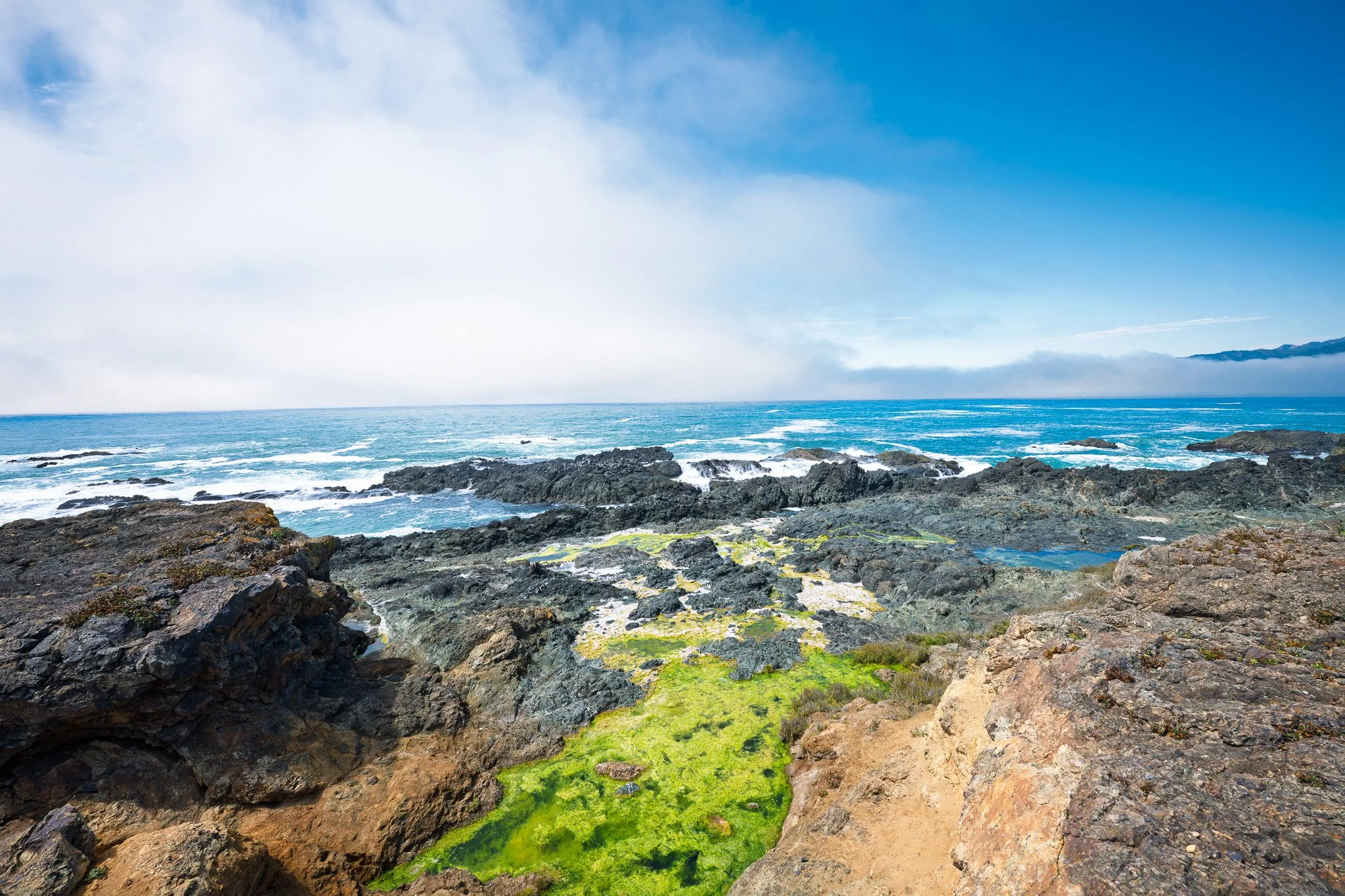 Rocky coastline with patches of green moss, ocean waves, and a mostly clear sky with some clouds.