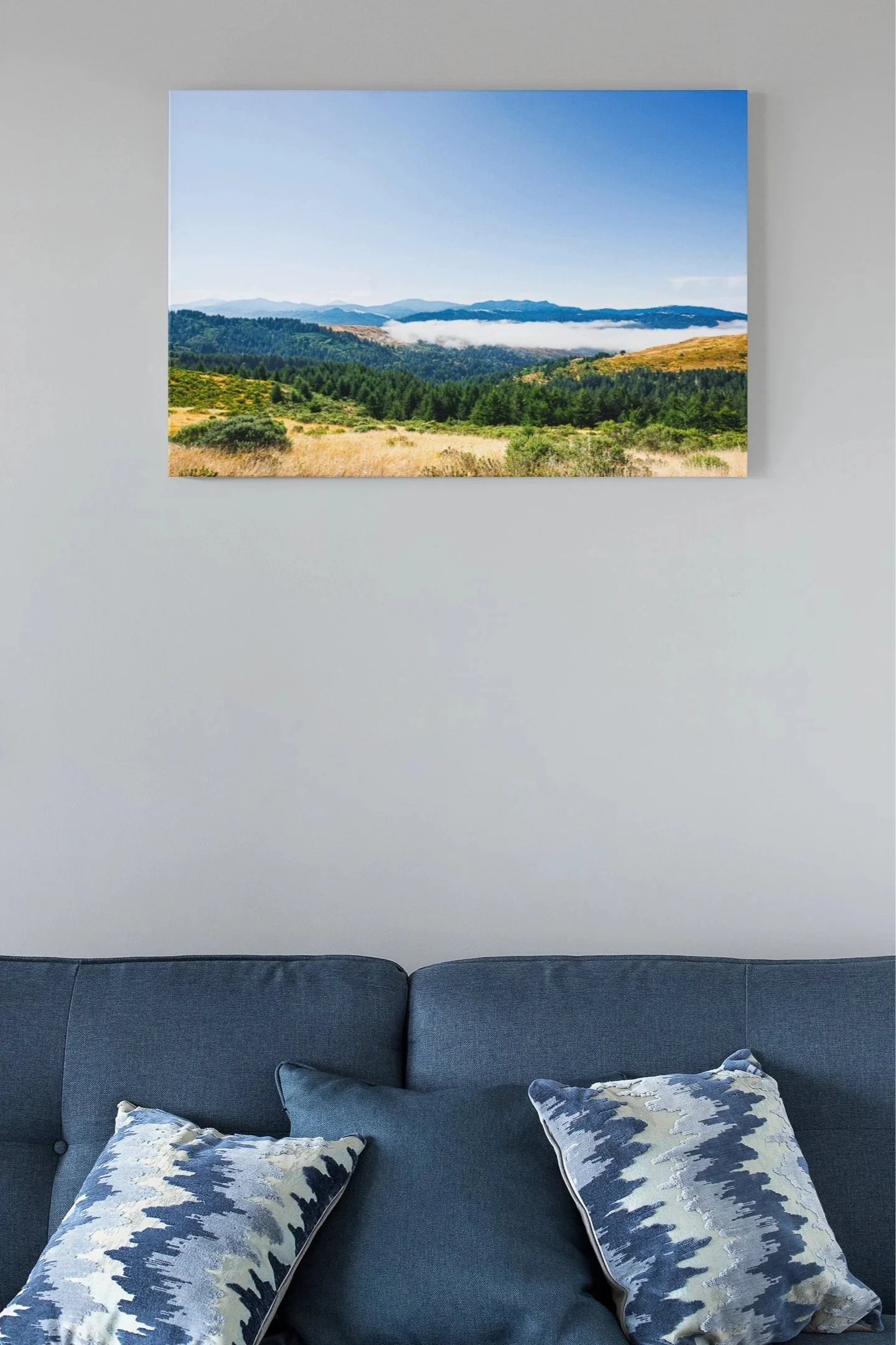 A landscape photograph of rolling hills, green trees, and a blue sky with clouds, hanging on a white wall above a blue sofa with decorative pillows.