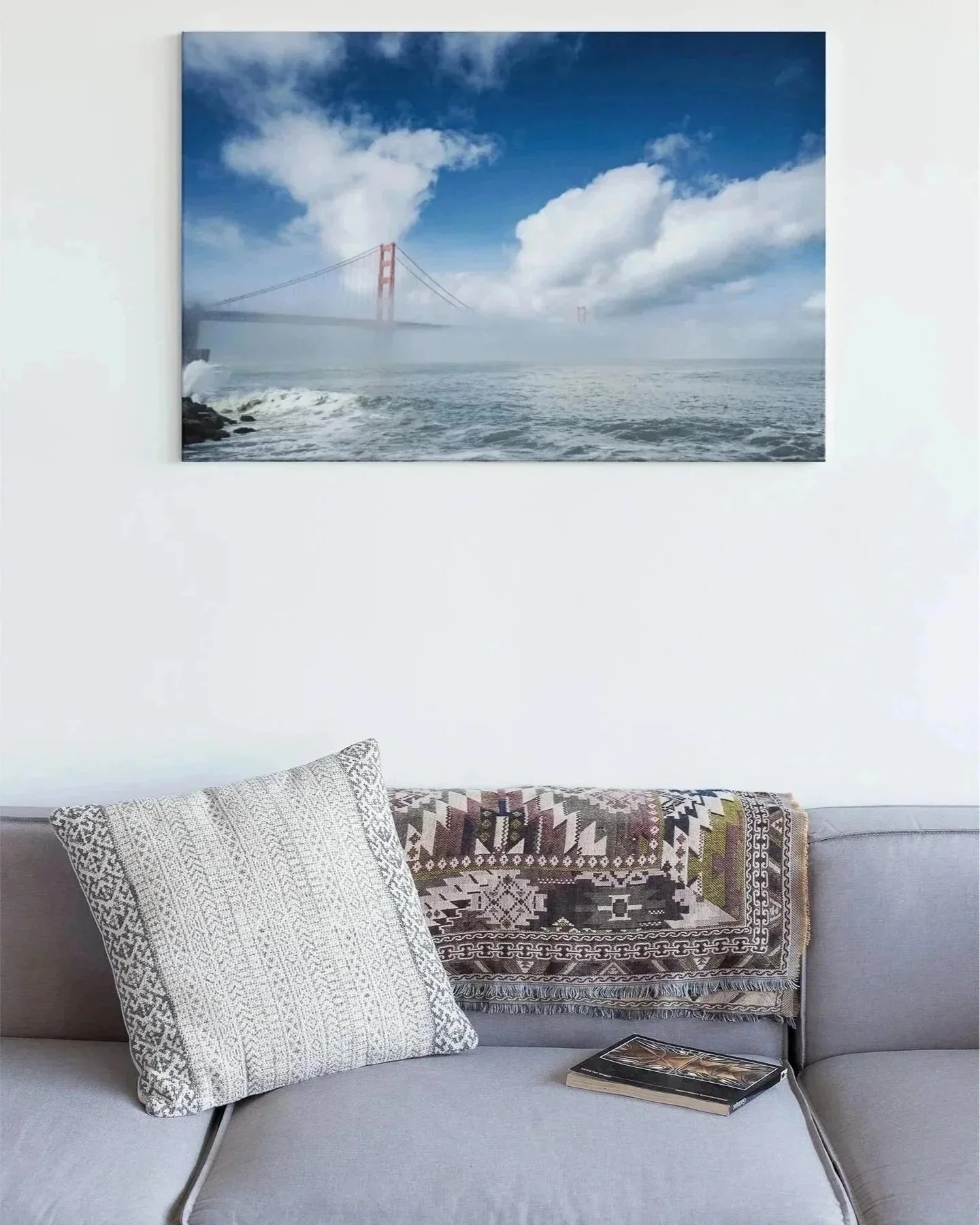 A living room with a light gray sofa, decorative pillow, and a patterned blanket. A book is placed on the sofa. Above, there's a large wall art depicting a foggy view of the Golden Gate Bridge over the water under a partly cloudy sky.