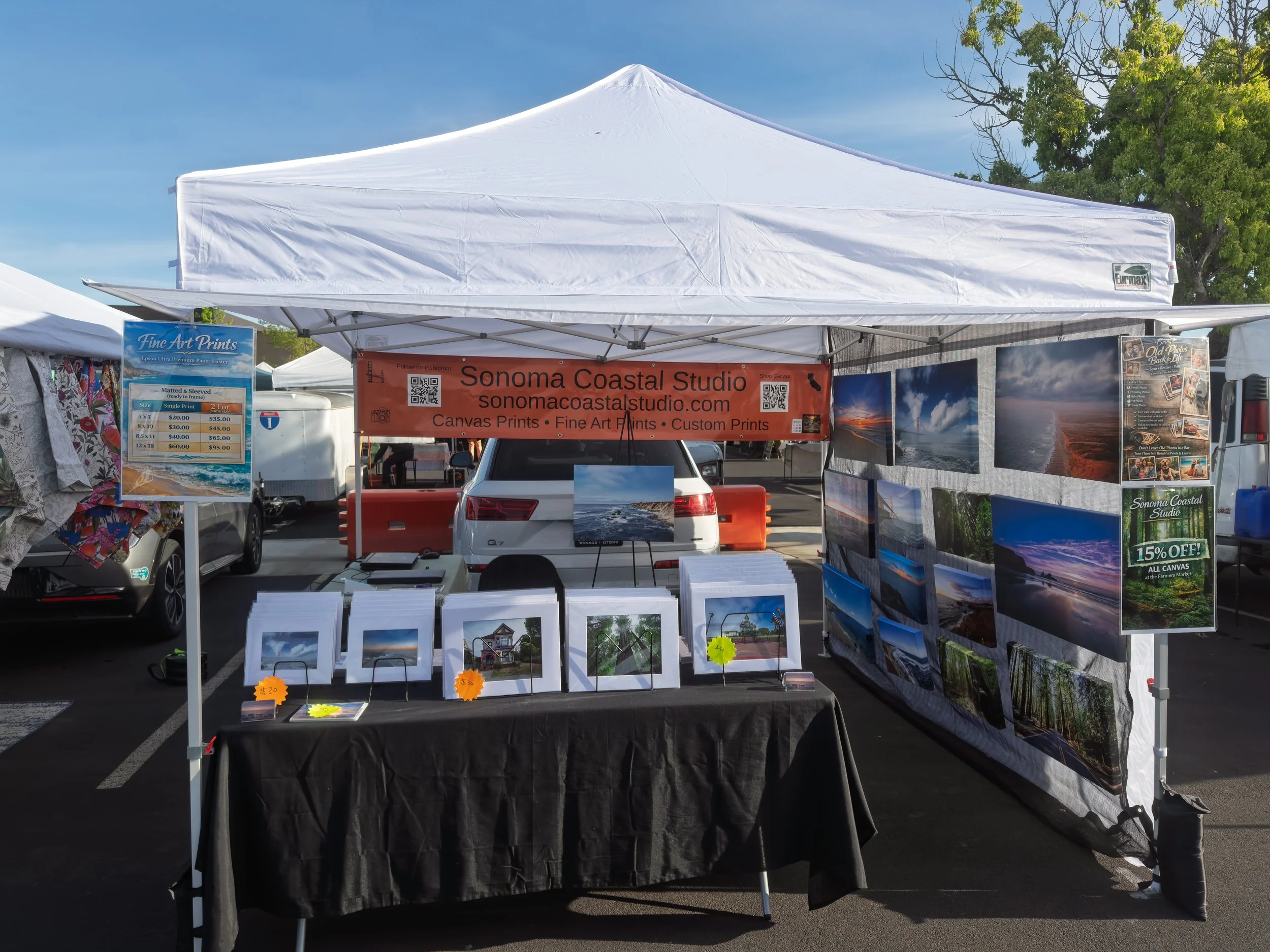 An outdoor art booth selling canvas prints at a market event, featuring landscape photographs of beaches, forests, and sunsets, with prices displayed on signs.