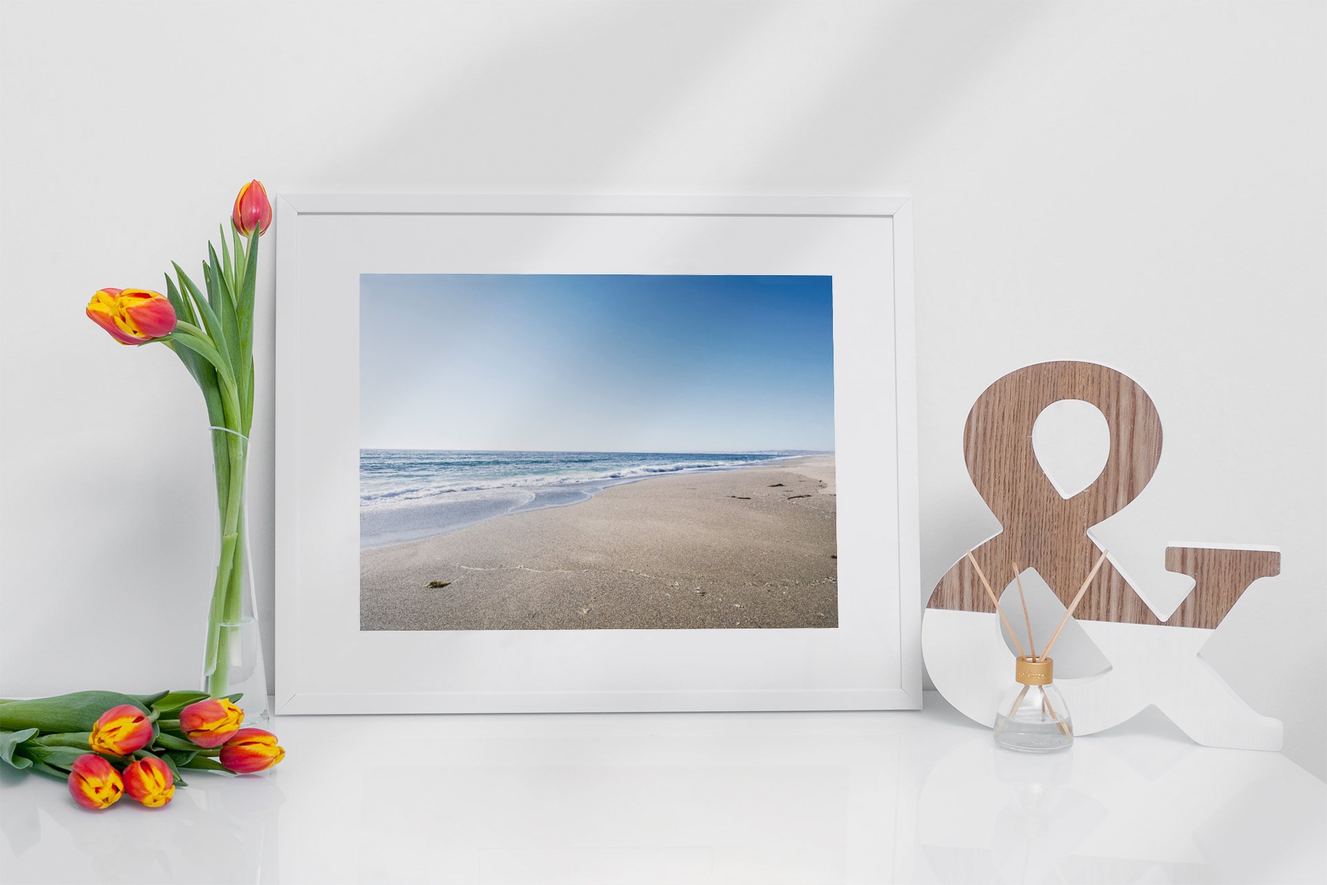 Decorative white surface with a framed beach photograph, a clear vase with pink and yellow tulips, and a wooden ampersand symbol with a small glass container holding reed diffusers.