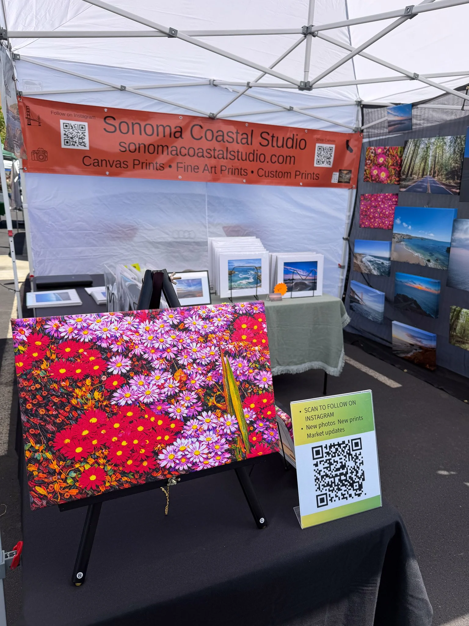 An outdoor art booth displaying a vibrant floral painting of red, pink, and purple flowers on a black background. Behind the painting, there are several framed landscape photographs of beaches, ocean, and nature scenes. A red banner reads 'Sonoma Coastal Studio' with website and services listed, and QR codes are attached for social media and contact information. A sign with a QR code at the front invites visitors to follow on Instagram and check for new photos and prints.