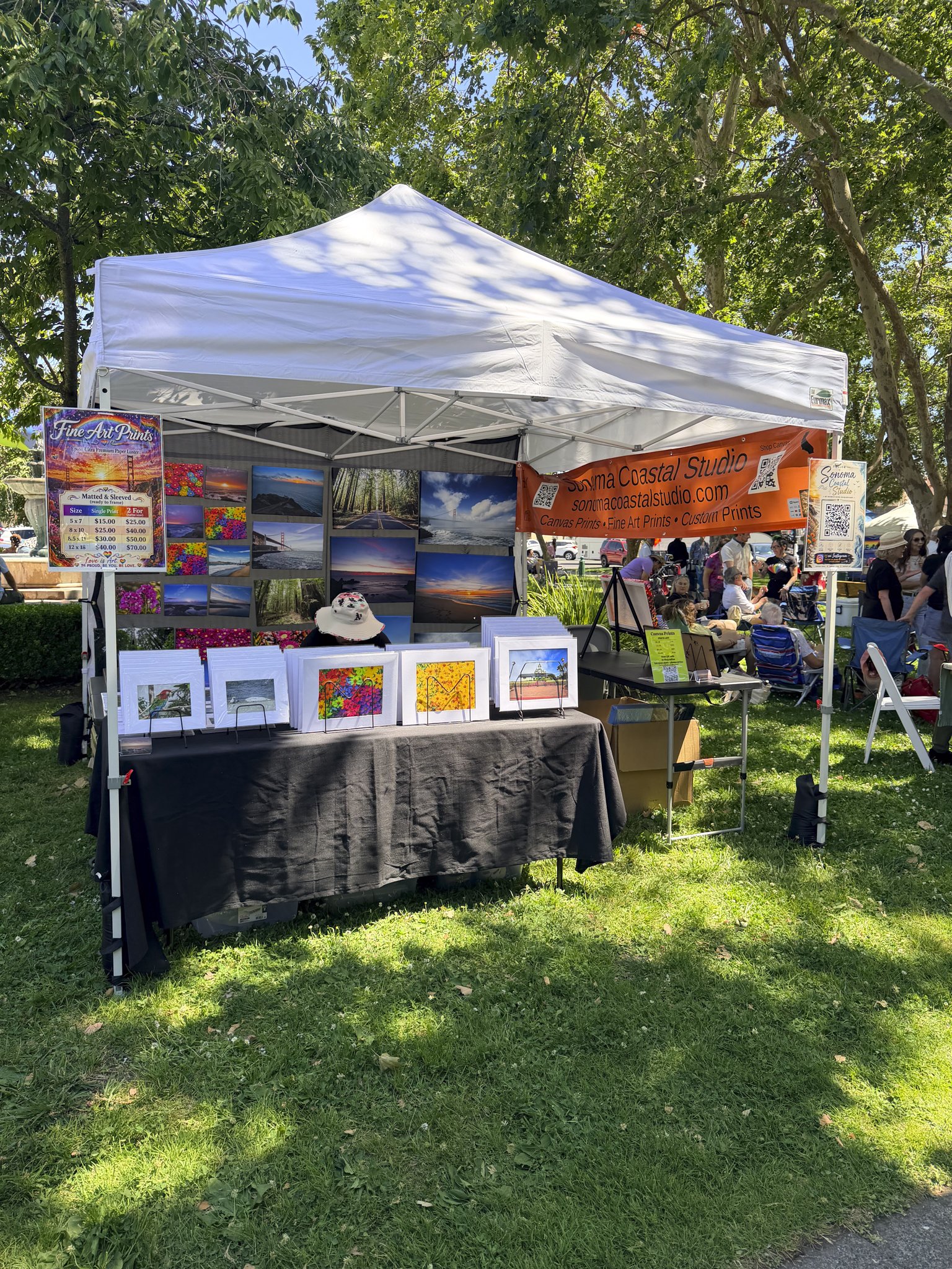 A white canopy tent at an outdoor art fair, with colorful framed paintings and prints displayed on tables and walls. There are people seated and walking around in the background.