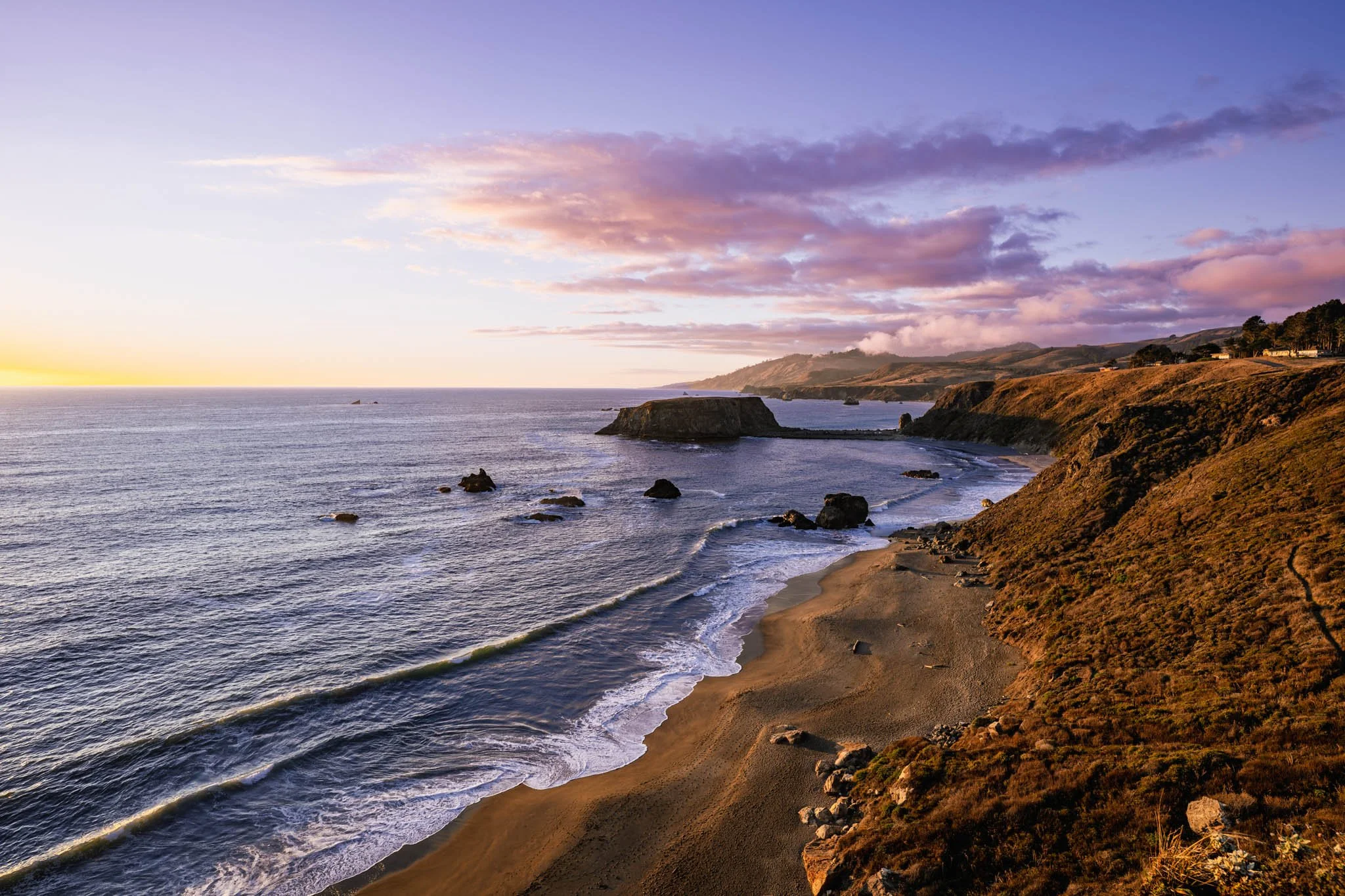 Sunset over the ocean along a rugged coastline with cliffs, rocky outcroppings, and a sandy beach, under a sky with scattered clouds