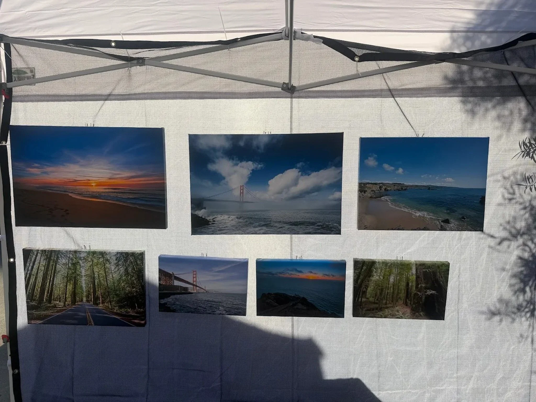 Set of nine landscape photographs displayed on a white fabric backdrop inside a canopy tent, featuring scenes of sunsets, beaches, forests, and the Golden Gate Bridge.
