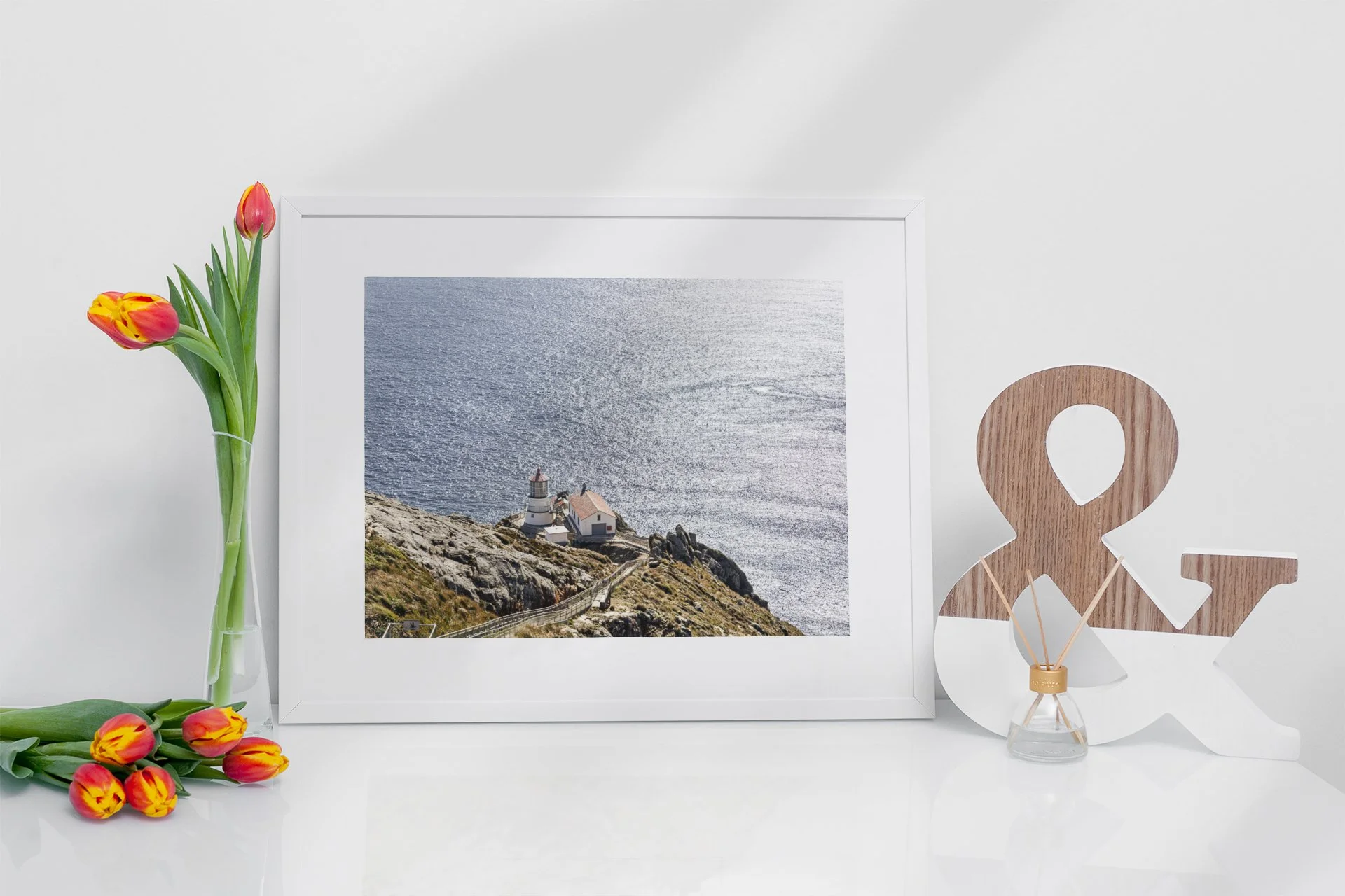 Decorative arrangement featuring a framed photo of a lighthouse and ocean, a vase with pink and yellow tulips, and a wooden ampersand sculpture on a white surface.