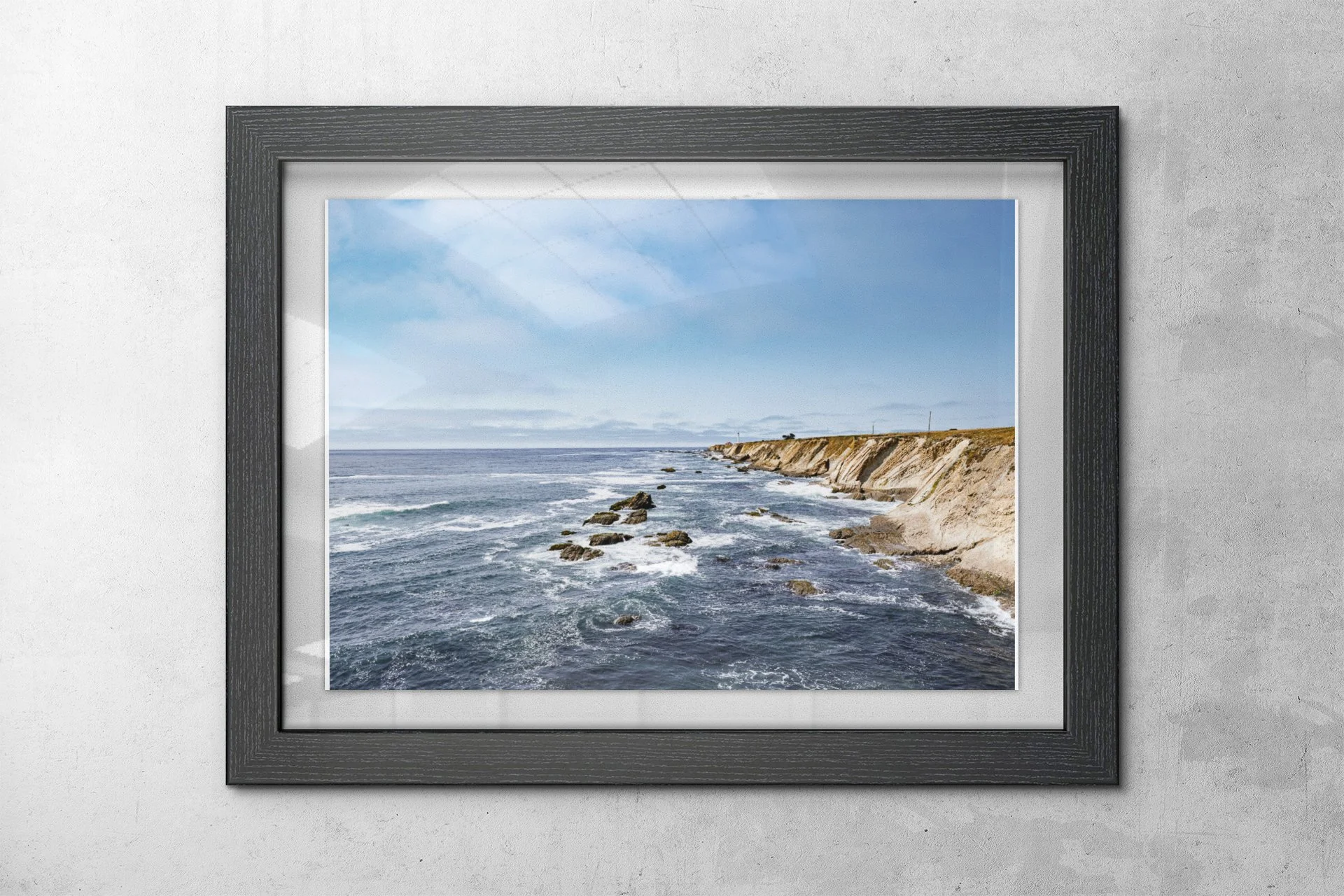 A framed photograph of a coastal view with waves crashing against rocks and a sandy shoreline, under a partly cloudy sky.