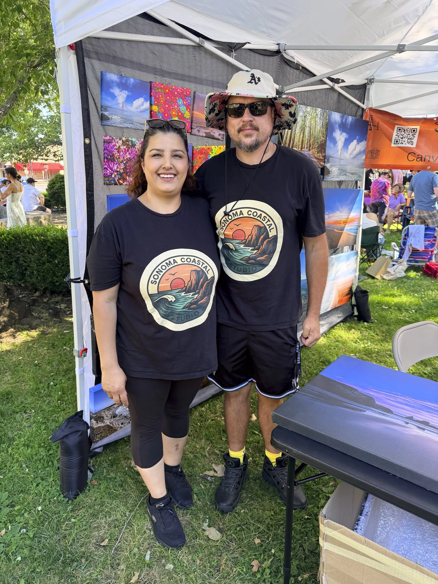 A woman and man stand together at an outdoor art fair, both wearing black T-shirts with a coastal sunset design. The woman has long brown hair, sunglasses on her head, and is smiling at the camera. The man has a beard, pink patterned hat, sunglasses, and is standing with his arm around the woman. Behind them is a display of landscape photographs, and other people are visible in the background.
