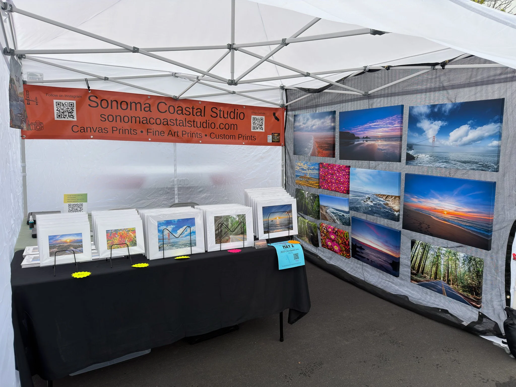 An art and photography booth at an outdoor event displaying framed landscape photographs on the walls and table. There are various scenery images, and a red banner reads "Sonoma Coastal Studio" with website URL and info about canvas prints and custom art.