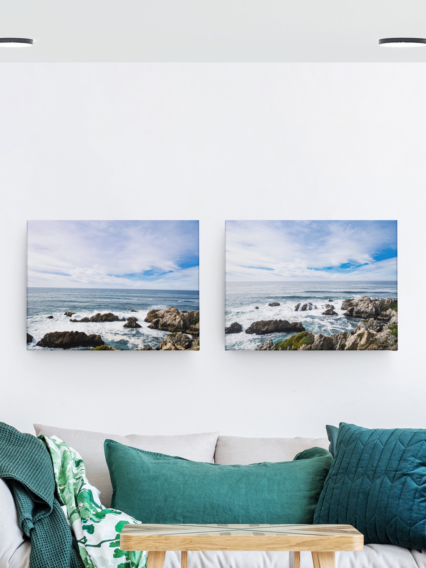 Two framed landscape photographs of ocean scenes hanging on a white wall over a beige couch with green and teal cushions.