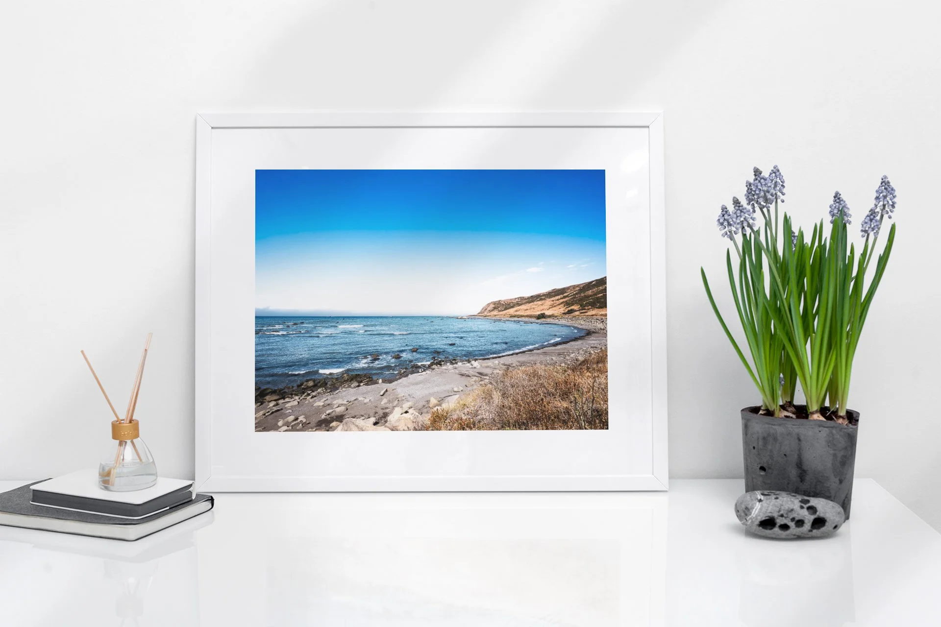 Framed landscape photograph of a beach with rocks, water, and hills on a white wall.
