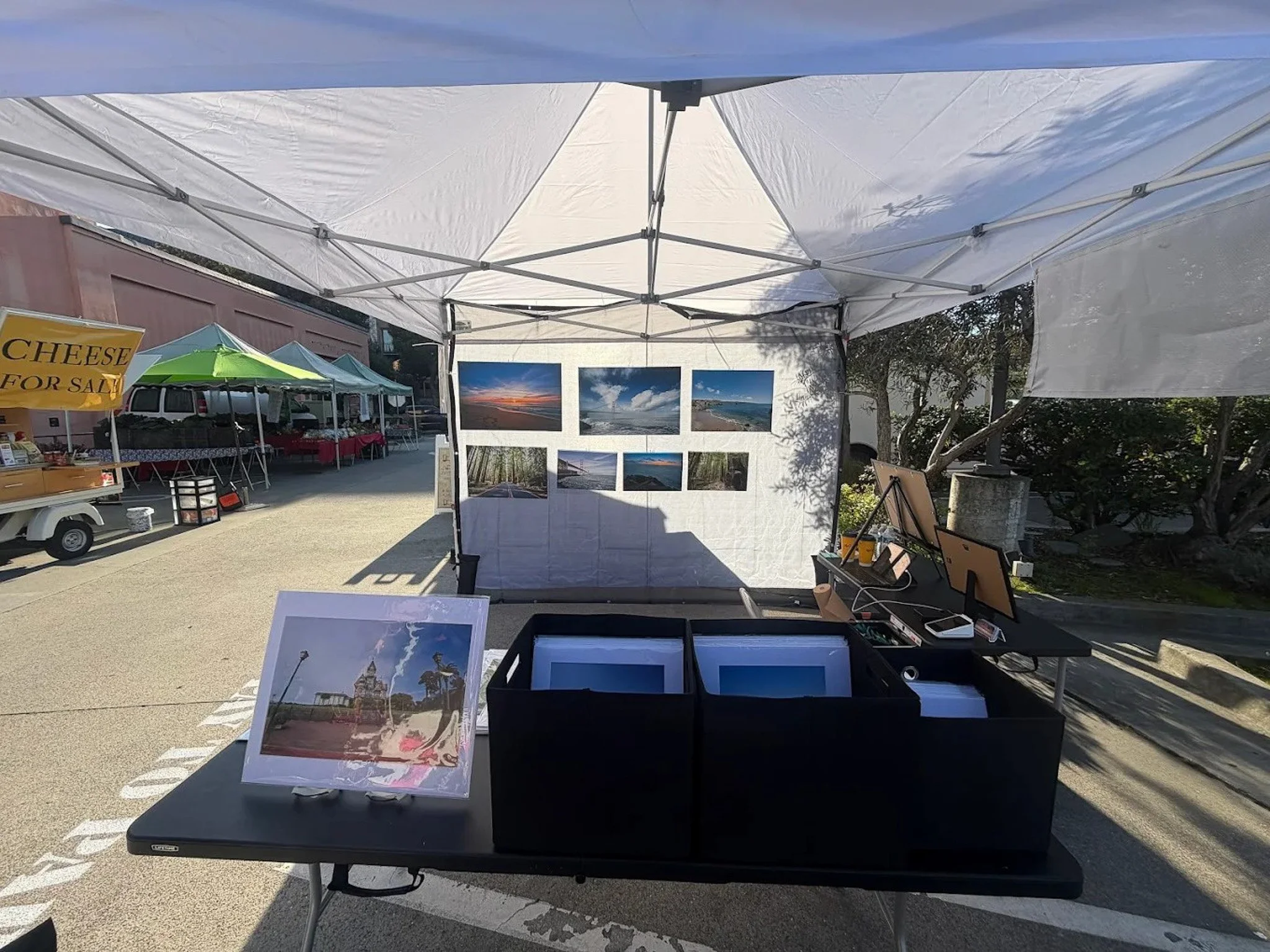 An outdoor art and photography booth under a white canopy, displaying landscape photographs on a back wall and a small framed photo on a table in front, at a market or fair.