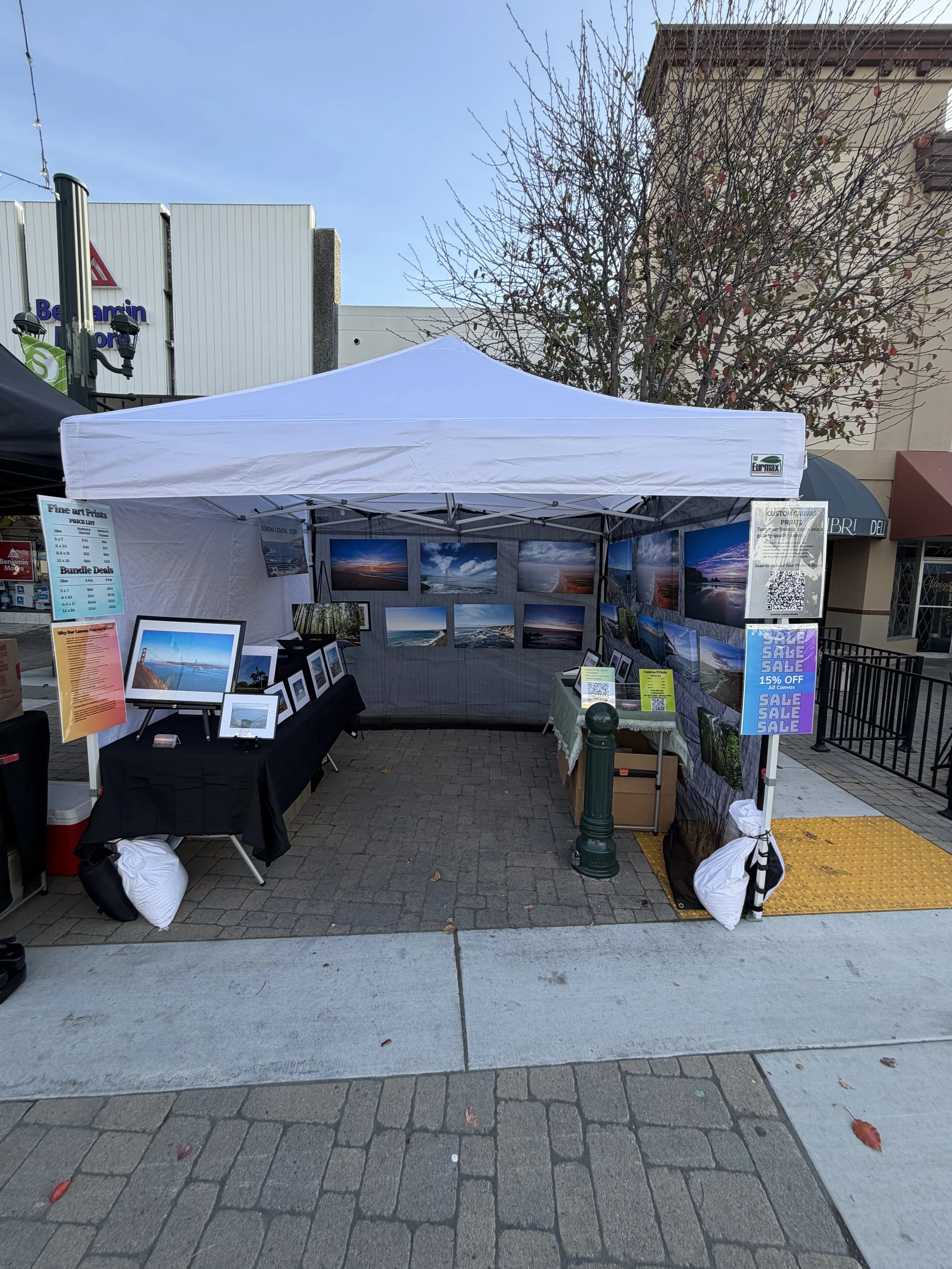 Street art vendor booth displaying landscape photographs for sale, with signs advertising discounts and pricing, set up on a sidewalk with a tree and buildings in the background.