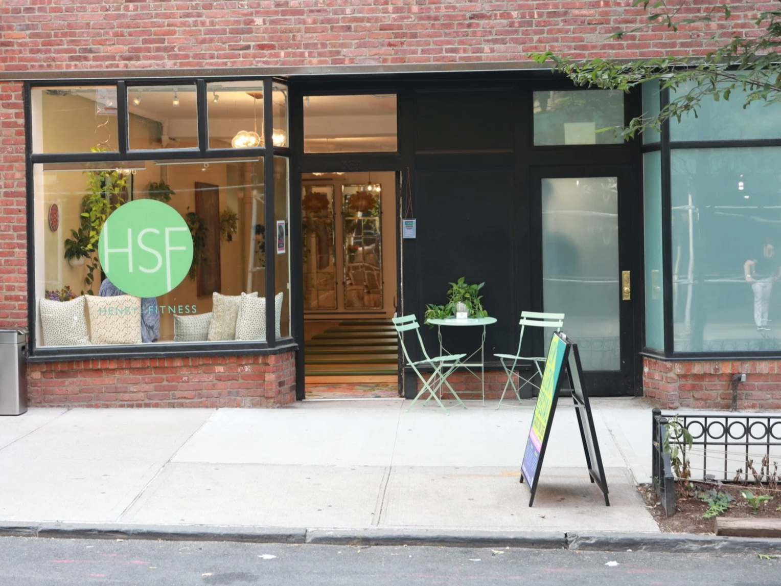 Exterior view of Henry Street Fitness on Henry Street in Cobble Hill Brooklyn. The photo depicts a green circular logo on the window, a small outdoor seating area with a white table and two chairs, potted plants, and an A-frame sign on the sidewalk.