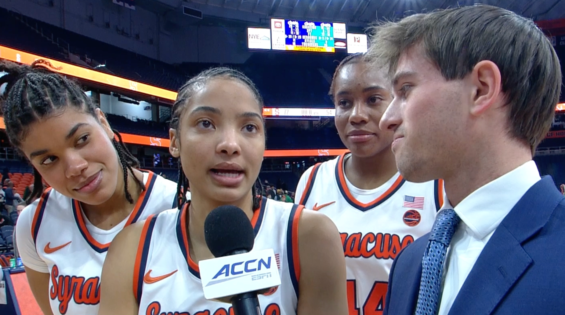 Four women in Syracuse women's basketball uniforms being interviewed by a man in a suit at a basketball stadium. One woman is speaking into a microphone labeled ACCN ESPN, while the others listen.
