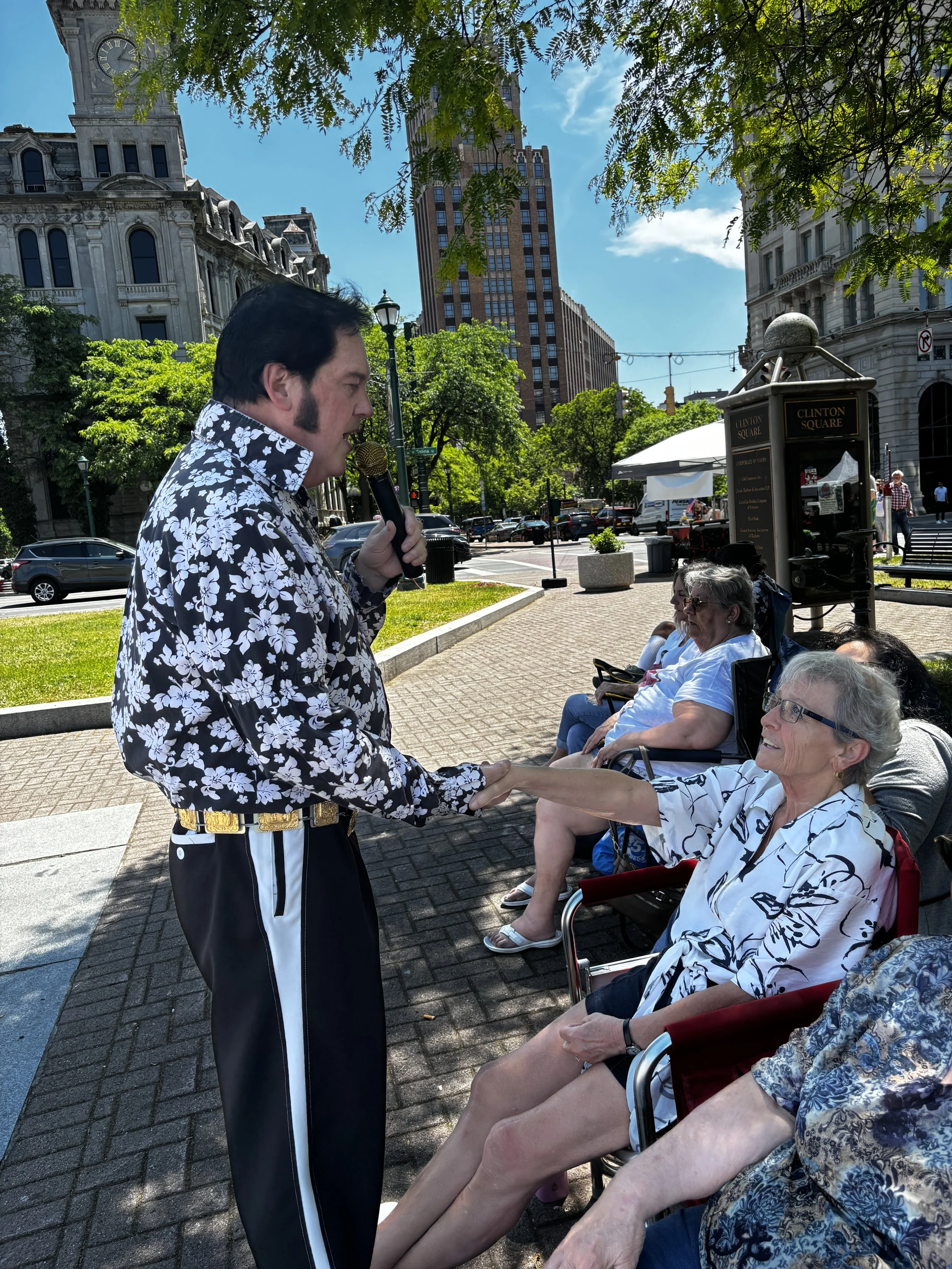 A man dressed in a floral shirt is holding hands with an elderly woman seated in a wheelchair in a park in Clinton Square, with other elderly women sitting nearby. There are tall buildings and trees in the background, and the sky is partly cloudy.