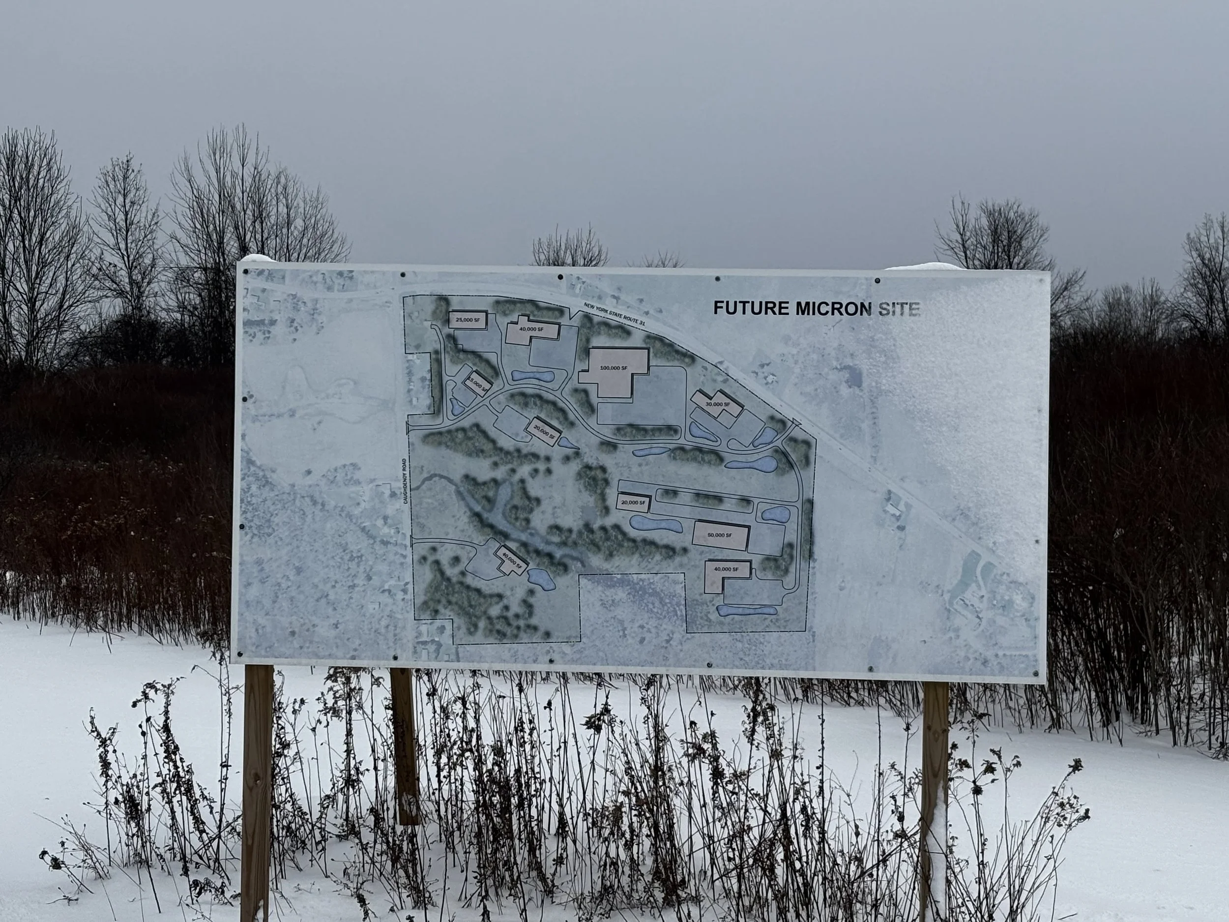 A site plan for a future MICRON facility displayed on a signboard outdoors in a snowy landscape with leafless trees in the background.
