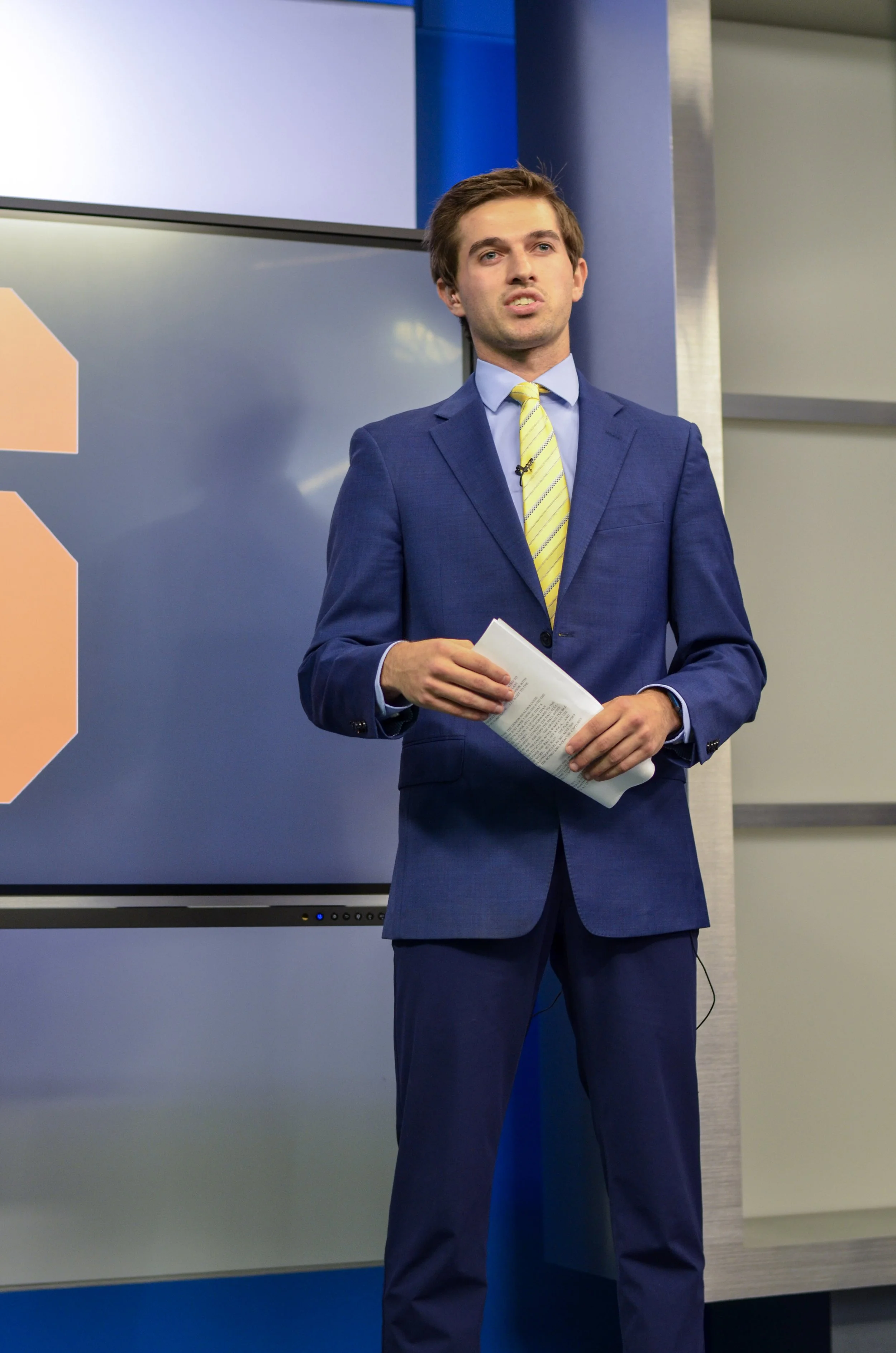 A man in a blue suit and yellow tie holding papers, standing in front of a presentation screen in a conference room.