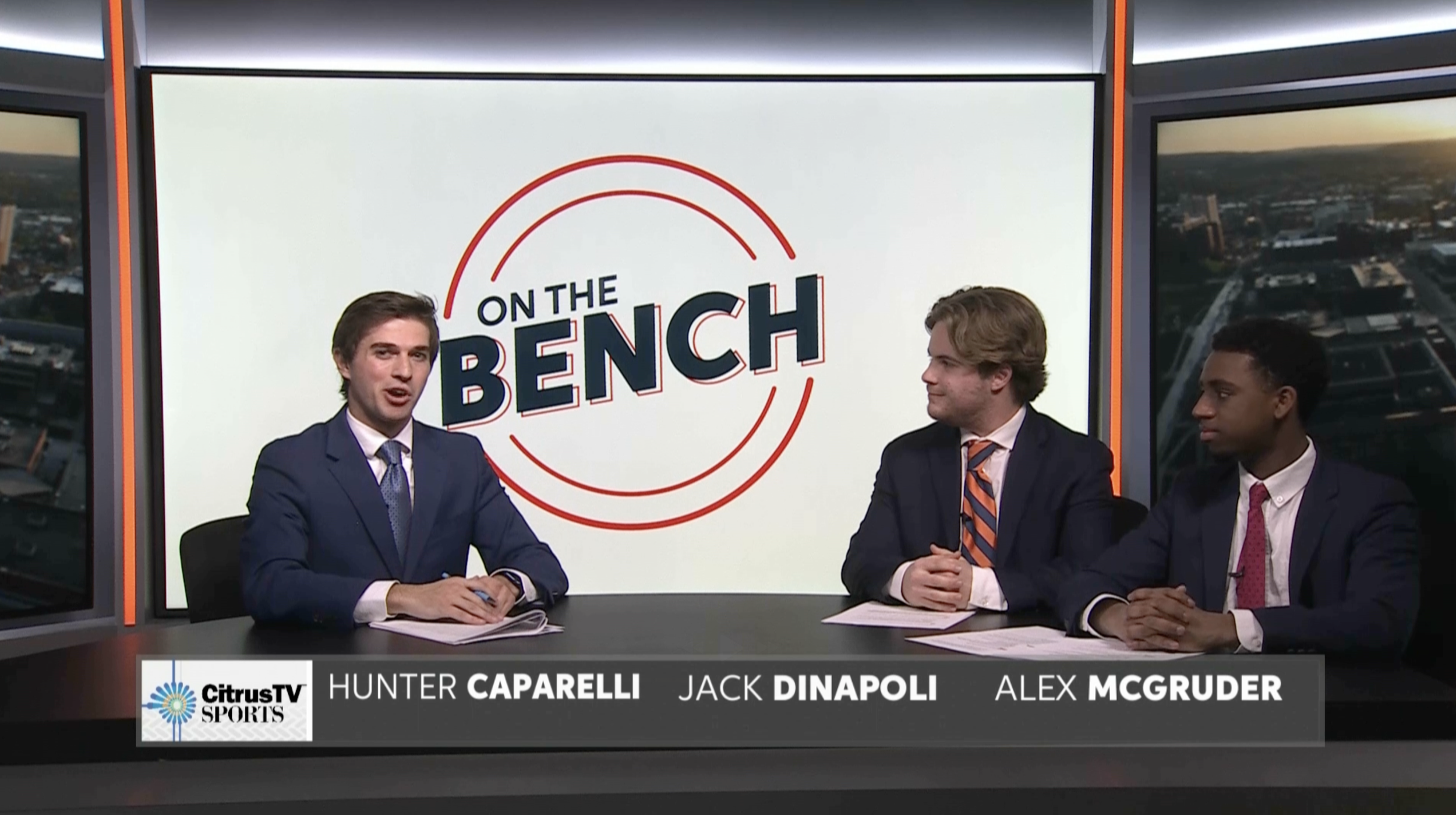 Three men in suits sitting at a news desk in a television studio with a large screen behind them displaying the text 'On the Bench'. The desk has a banner with the CitrusTV Sports logo and the names Hunter Caparelli, Jack Dinapoli, and Alex McGruder.