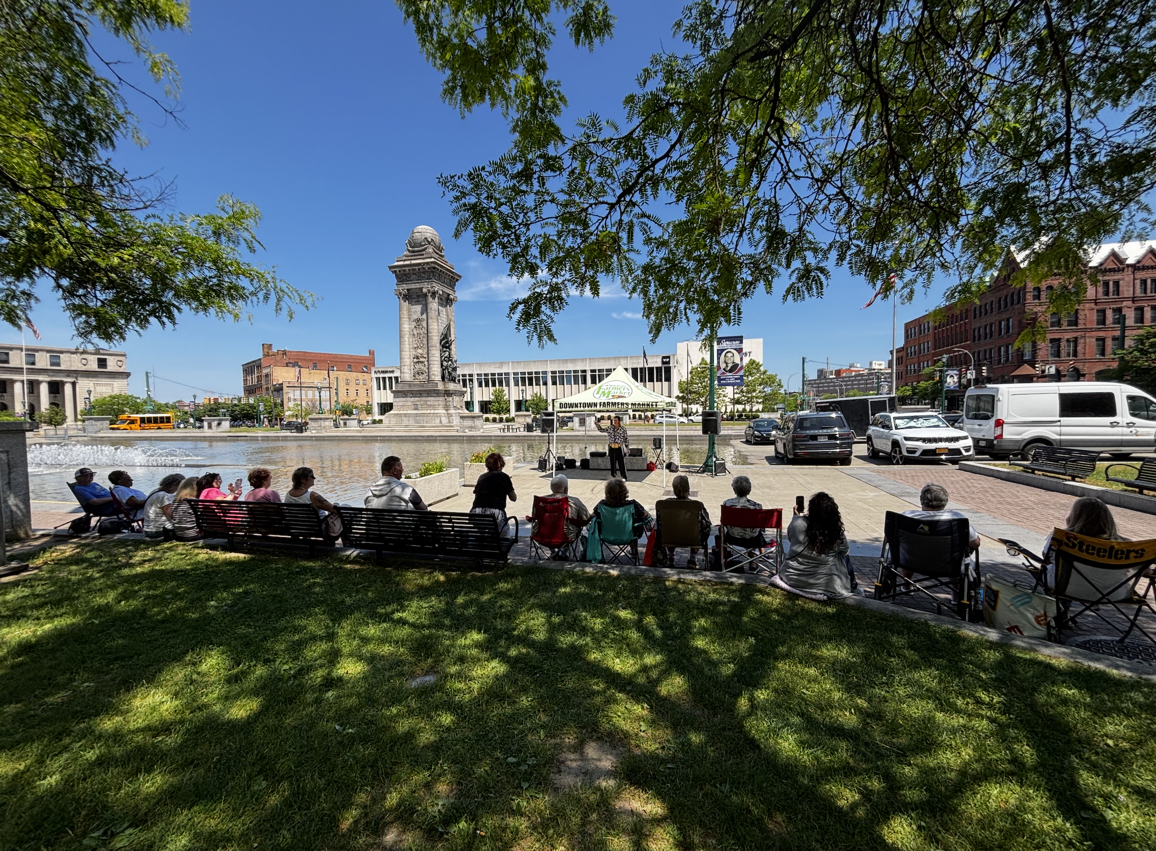 A park with a fountain, trees, and benches in an urban setting. People are sitting and standing, with vehicles visible on the street. A monument and some buildings are in the background.