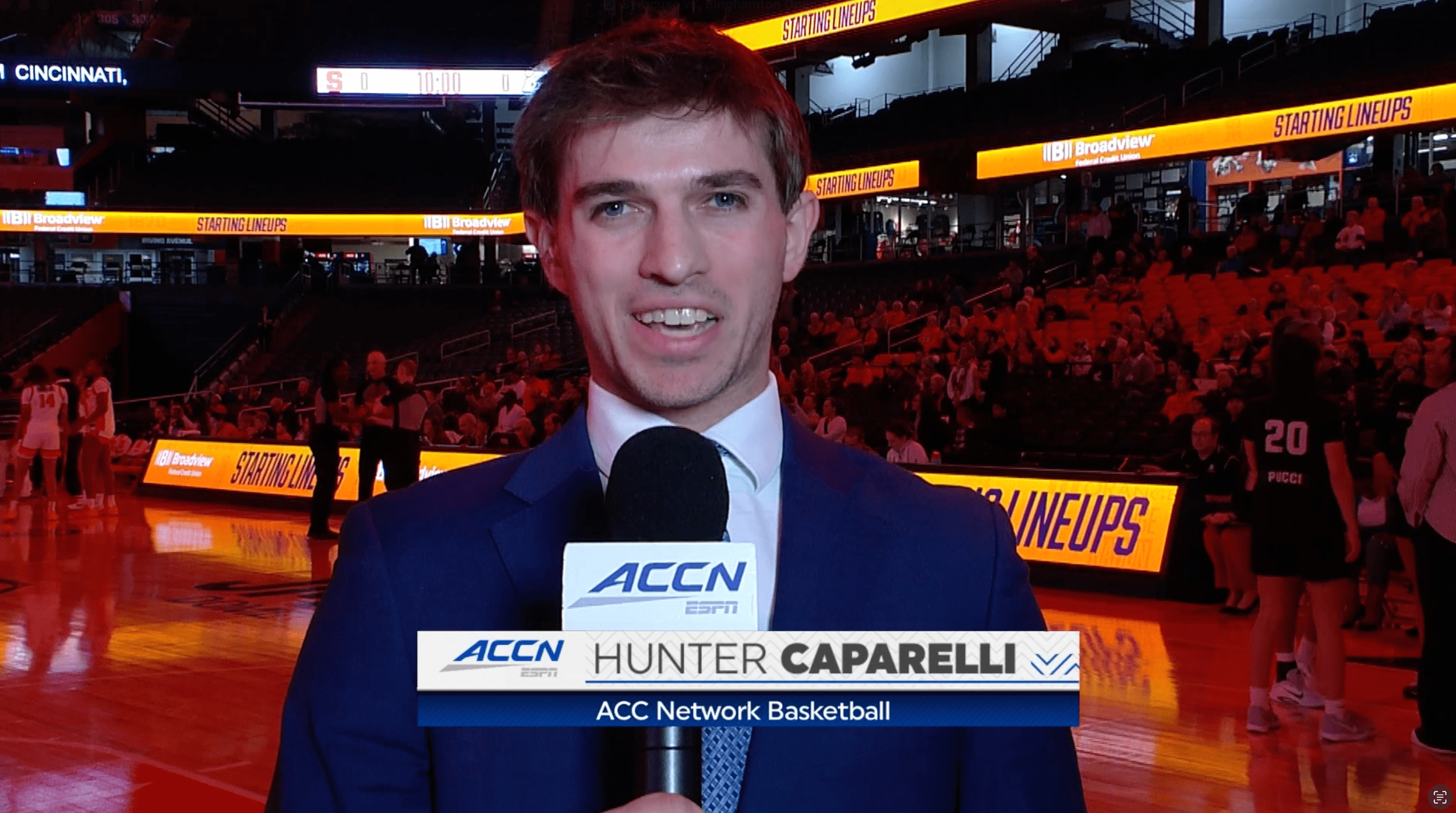 A male sports reporter holding a microphone with 'ACC Network ESPN' logo, standing on a basketball court with players and spectators in the background, during a basketball game event.