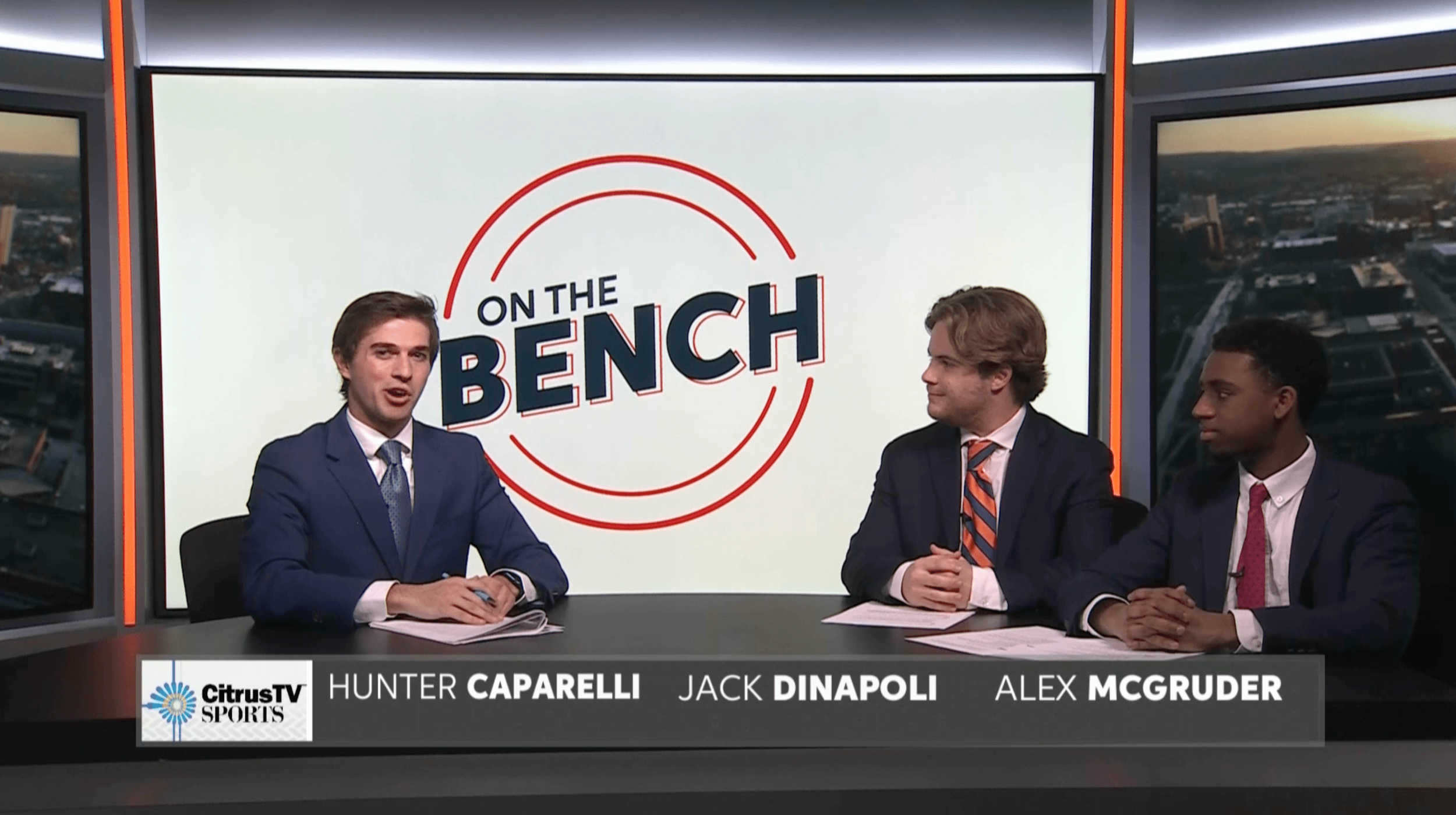 Three men in suits sitting at a news desk with a large screen behind them displaying the On the Bench logo. The screen shows a cityscape on the side, and there is a CitrusTV Sports logo with names Hunter Caparelli, Jack Dinapoli, and Alex McGruder at the bottom.