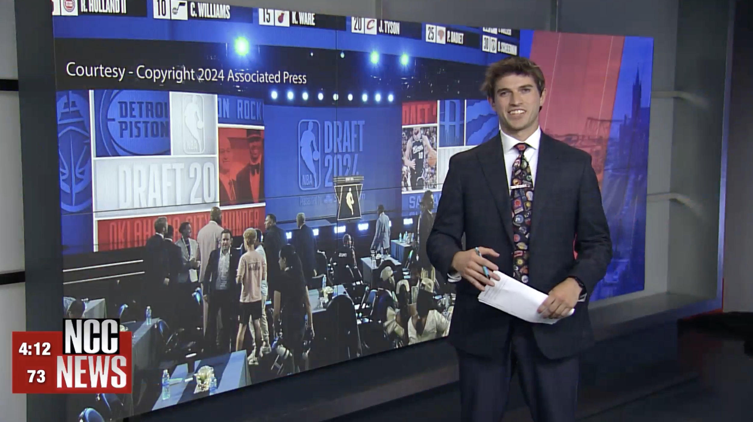 TV news studio with a male presenter standing in front of a large screen displaying NBA draft graphics and images of team logos and players, with people working at tables below on the studio floor.