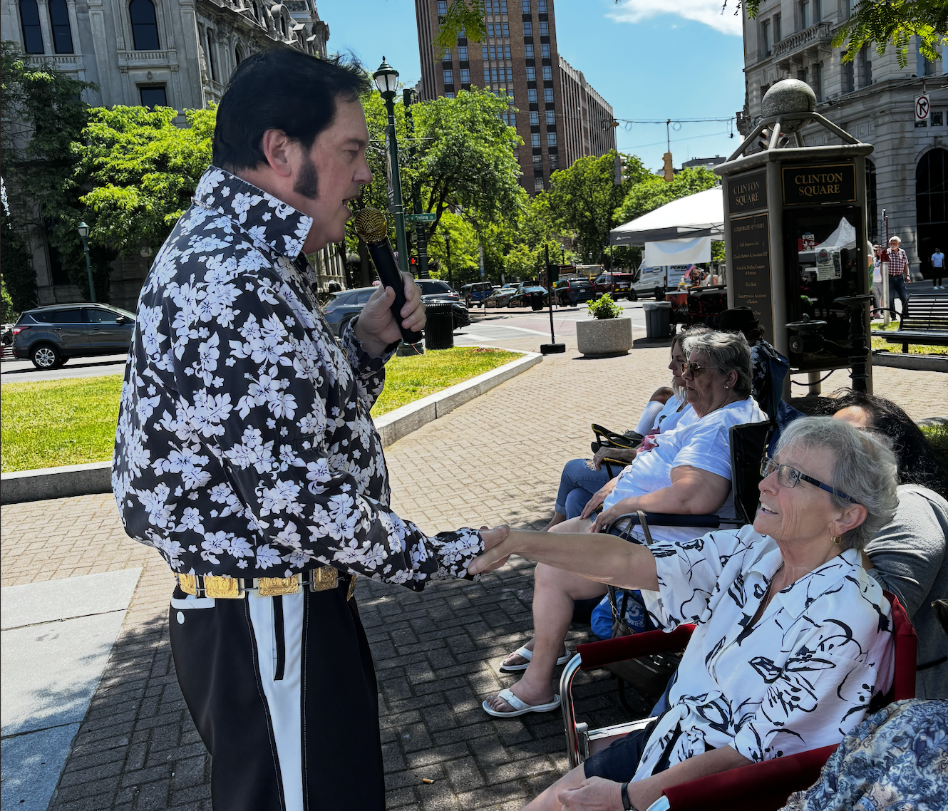 A man with dark hair, wearing a black and white floral shirt, holding a microphone and shaking hands with an elderly woman with gray hair and glasses, sitting in a red wheelchair, outdoors on a sunny day in an urban park with other seated elderly women and city buildings in the background.