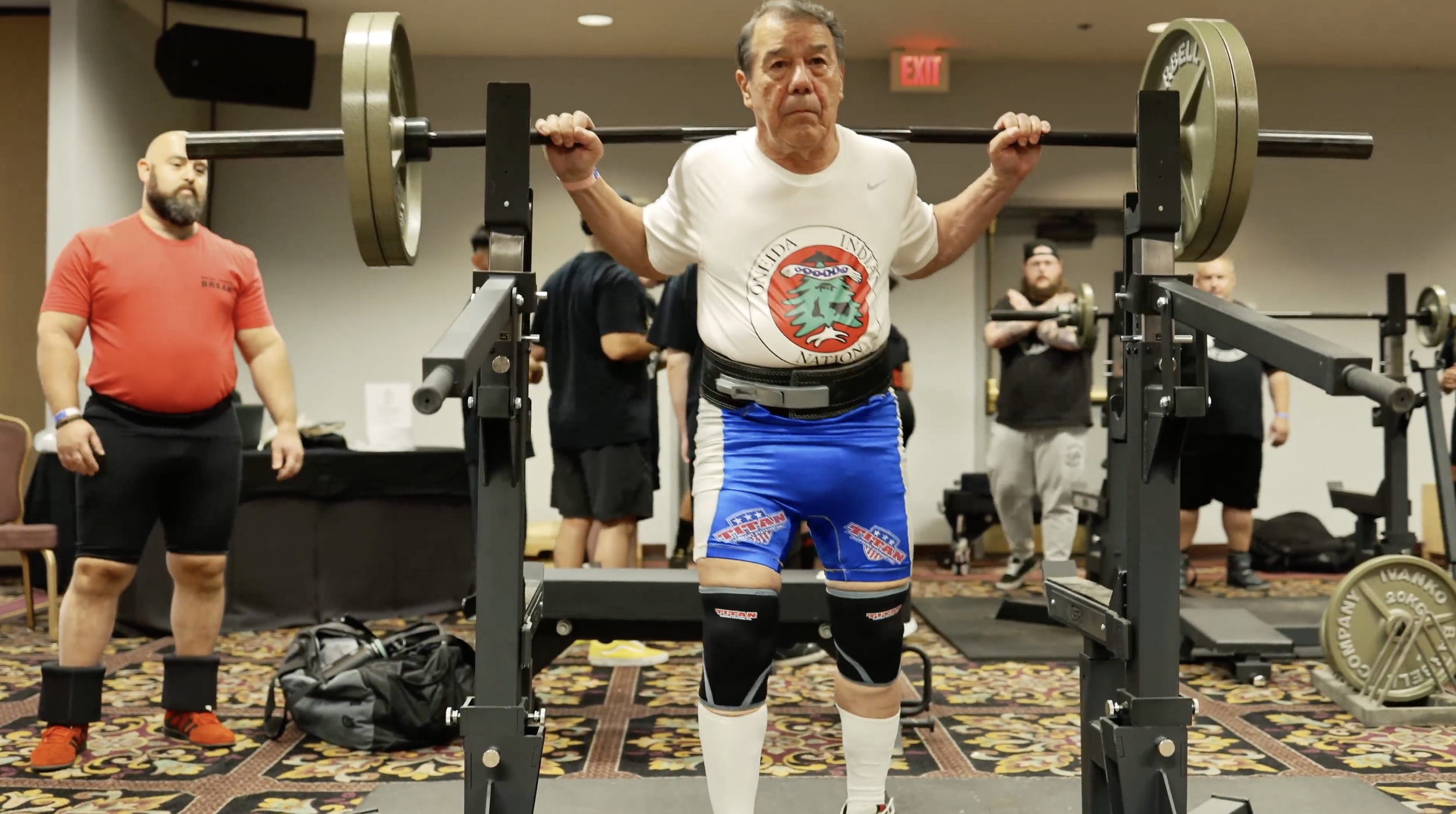 An elderly man with gray hair lifting a barbell with large weight plates on each end, standing on a lifting platform. He is wearing a white T-shirt with a logo, blue shorts, and knee-high socks. Several people are in the background, observing, including a man with a beard in a red T-shirt and black shorts.
