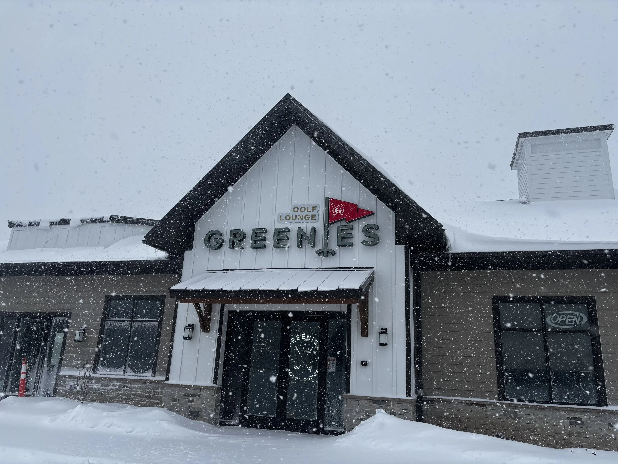Snow-covered building with a sign that says GOLF LOUNGE GREENS, with a red flag and snow falling.