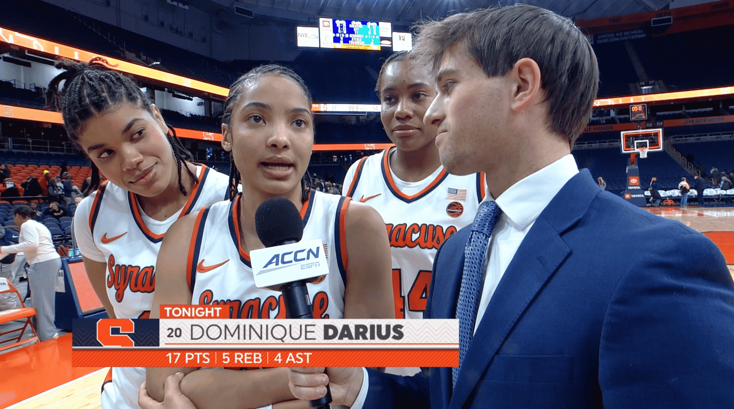 Three female basketball players from Syracuse University being interviewed by a male sports reporter after a game, inside a basketball arena.