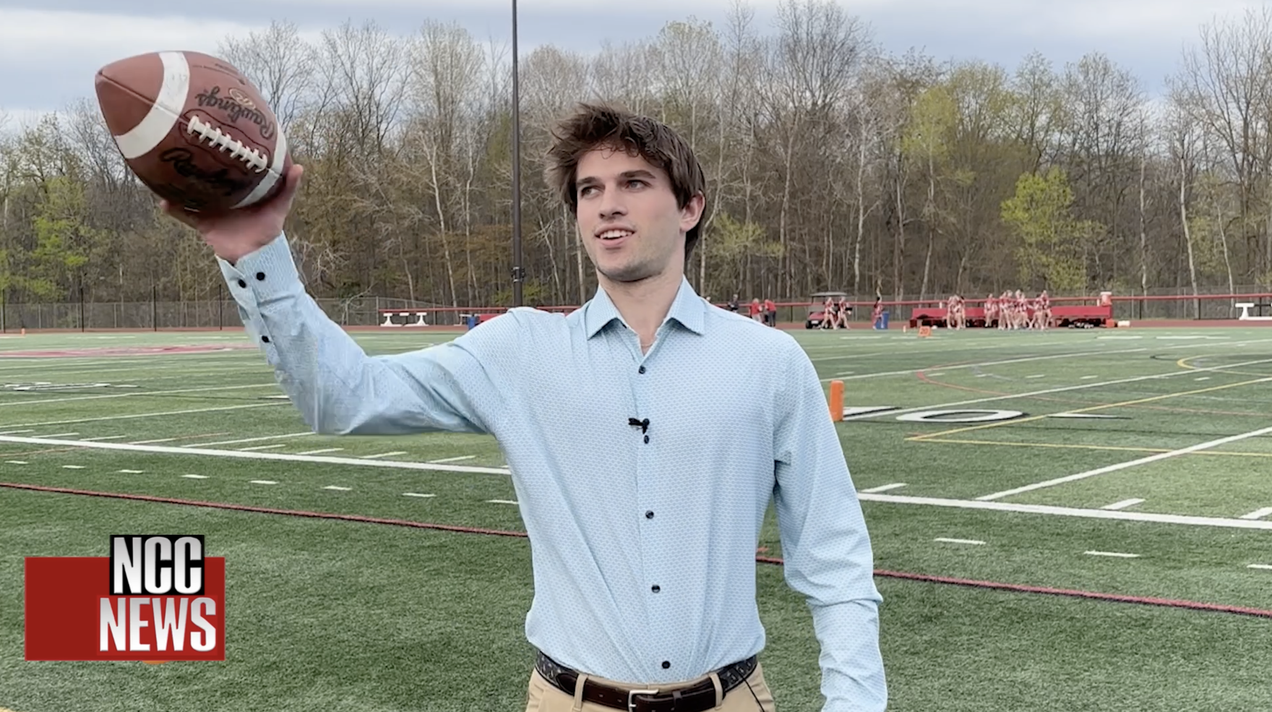 A young man in a light blue dress shirt holds a football in his right hand on a school football field with bare trees in the background. There is a red fence and a group of people in the distance. The image includes the 'NCC News' logo in the bottom left corner.