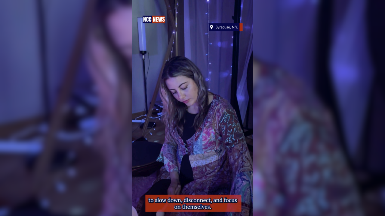 A woman sitting with her eyes closed, in a dimly lit room with blue and purple lighting, decorated with string lights, appearing to meditate or reflect, with television captions discussing slowing down, disconnecting, and focusing on themselves.