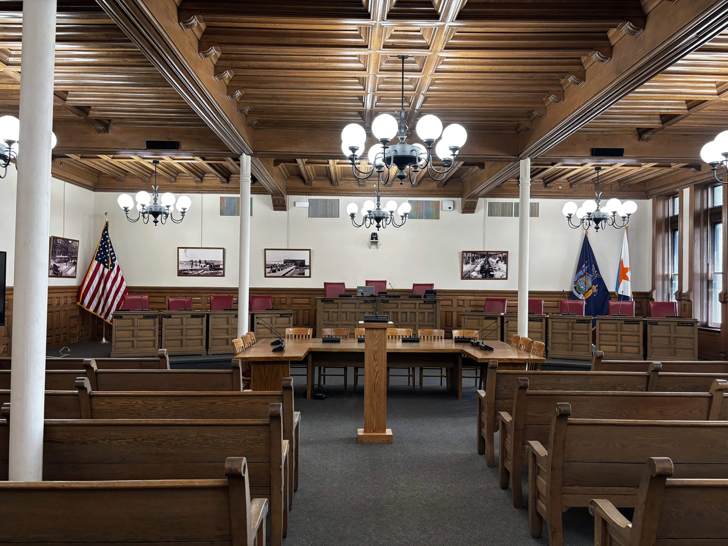 A formal government or courtroom chamber with a wooden ceiling, chandeliers, framed photographs, flags, rows of chairs, and a central wooden podium.