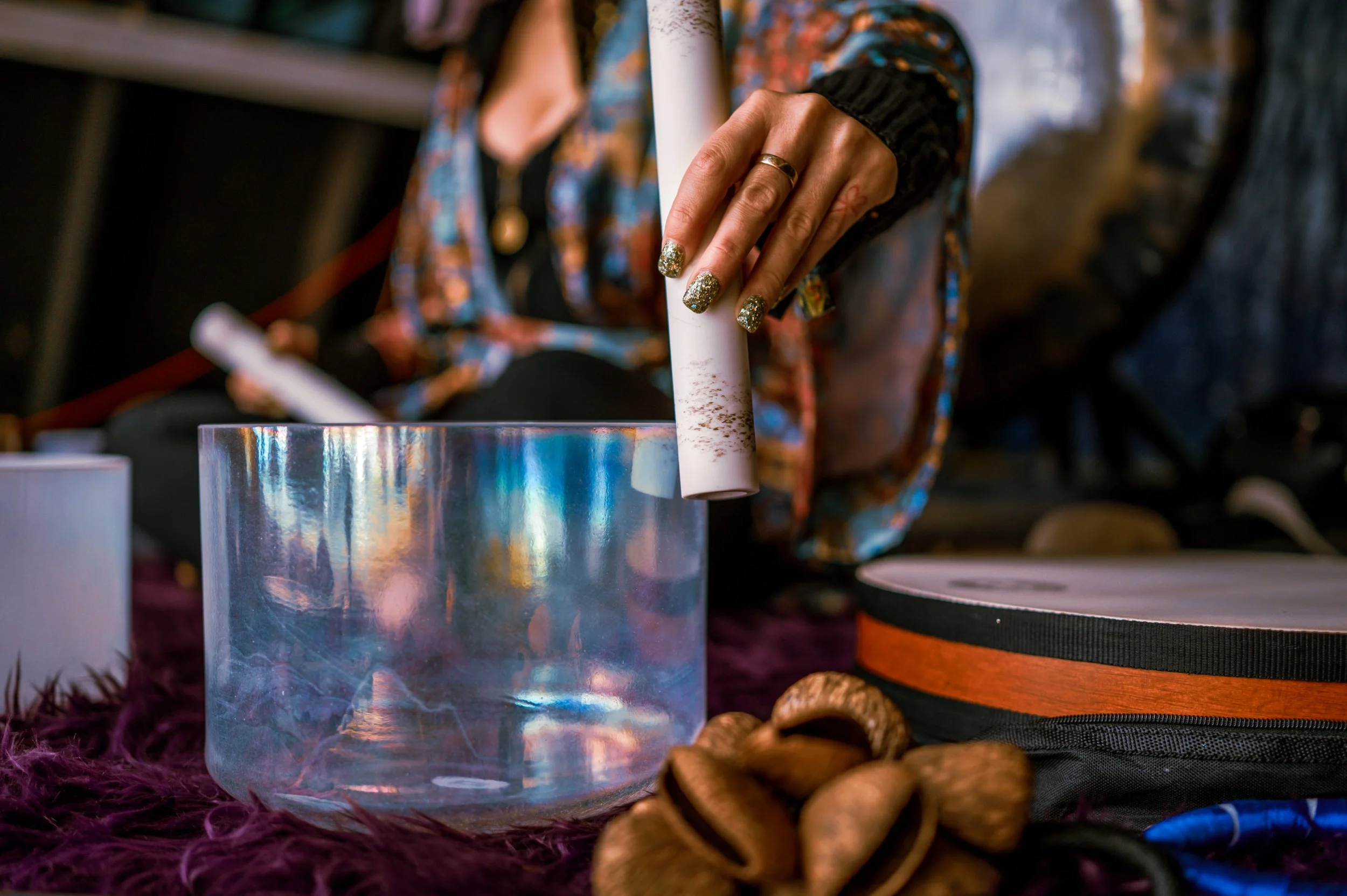 A person with glittery nails and rings is placing a rolled piece of white paper or cardboard into an iridescent, reflective container on a purple furry surface, with various objects including shells and a speaker surrounding it.
