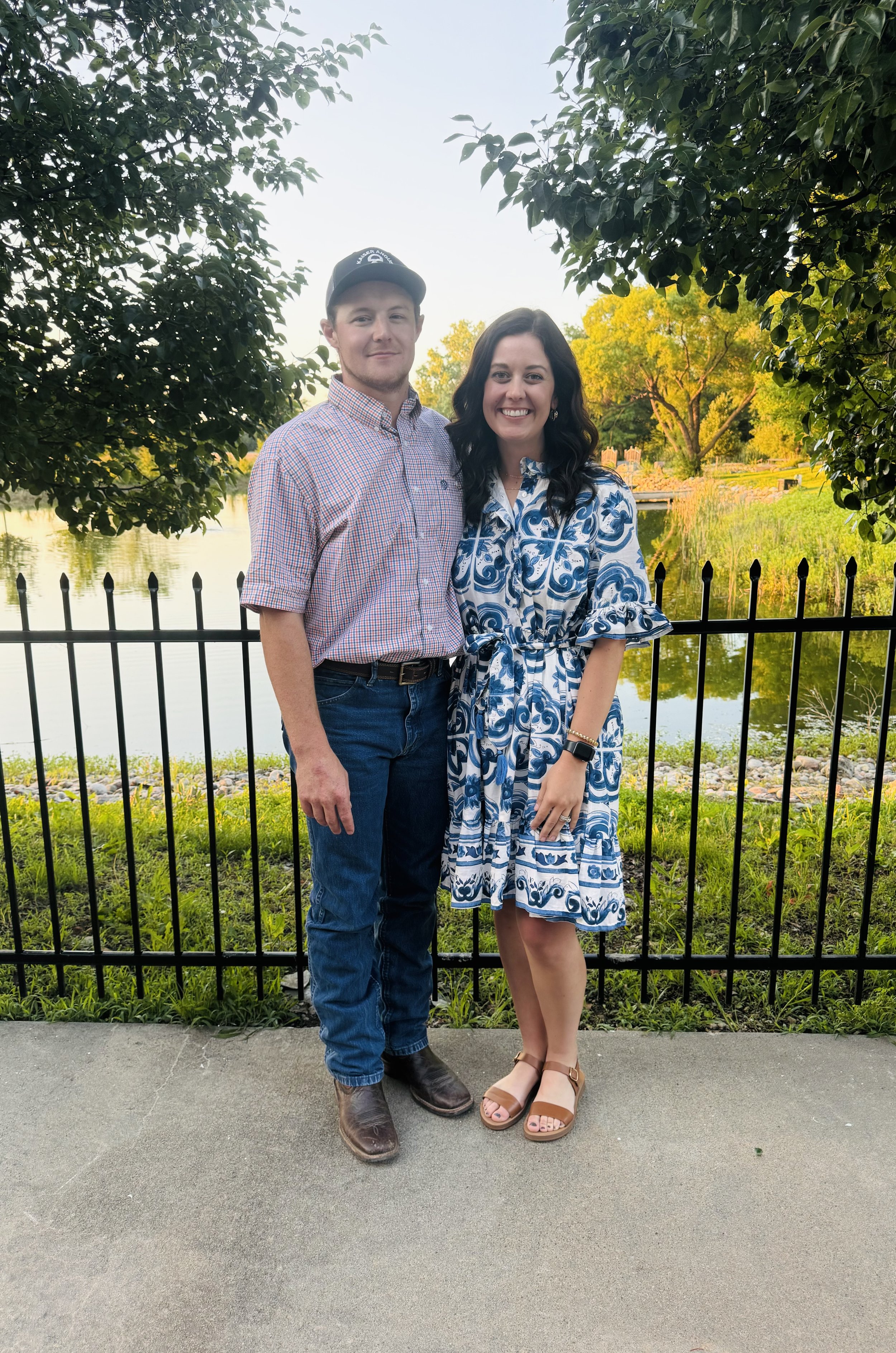 A man and woman standing by a black fence near a pond with trees in the background, smiling for the camera.