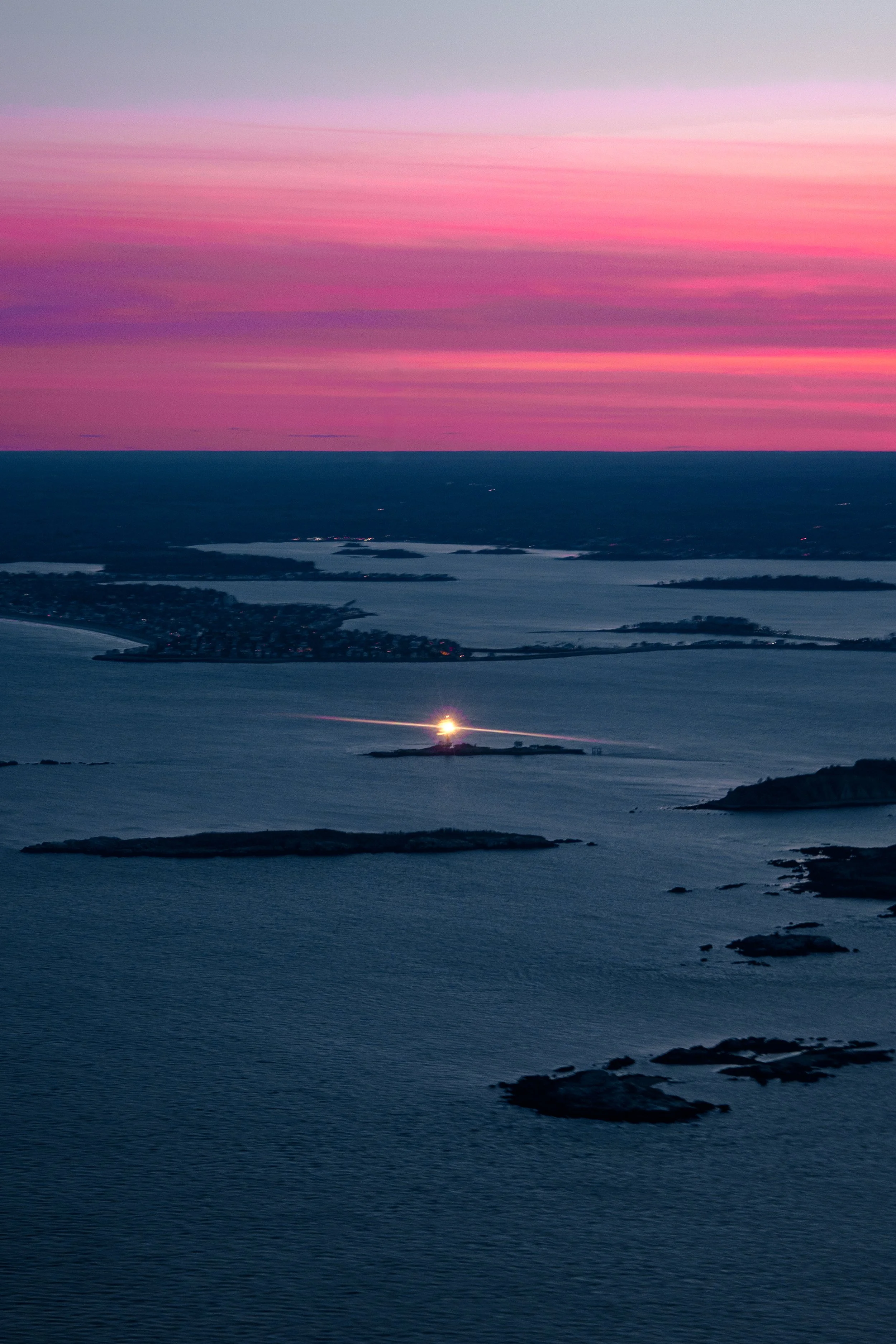 Aerial view of a body of water with islands at sunset, pink and purple hues in the sky, and a bright light on one of the islands reflecting on the water.