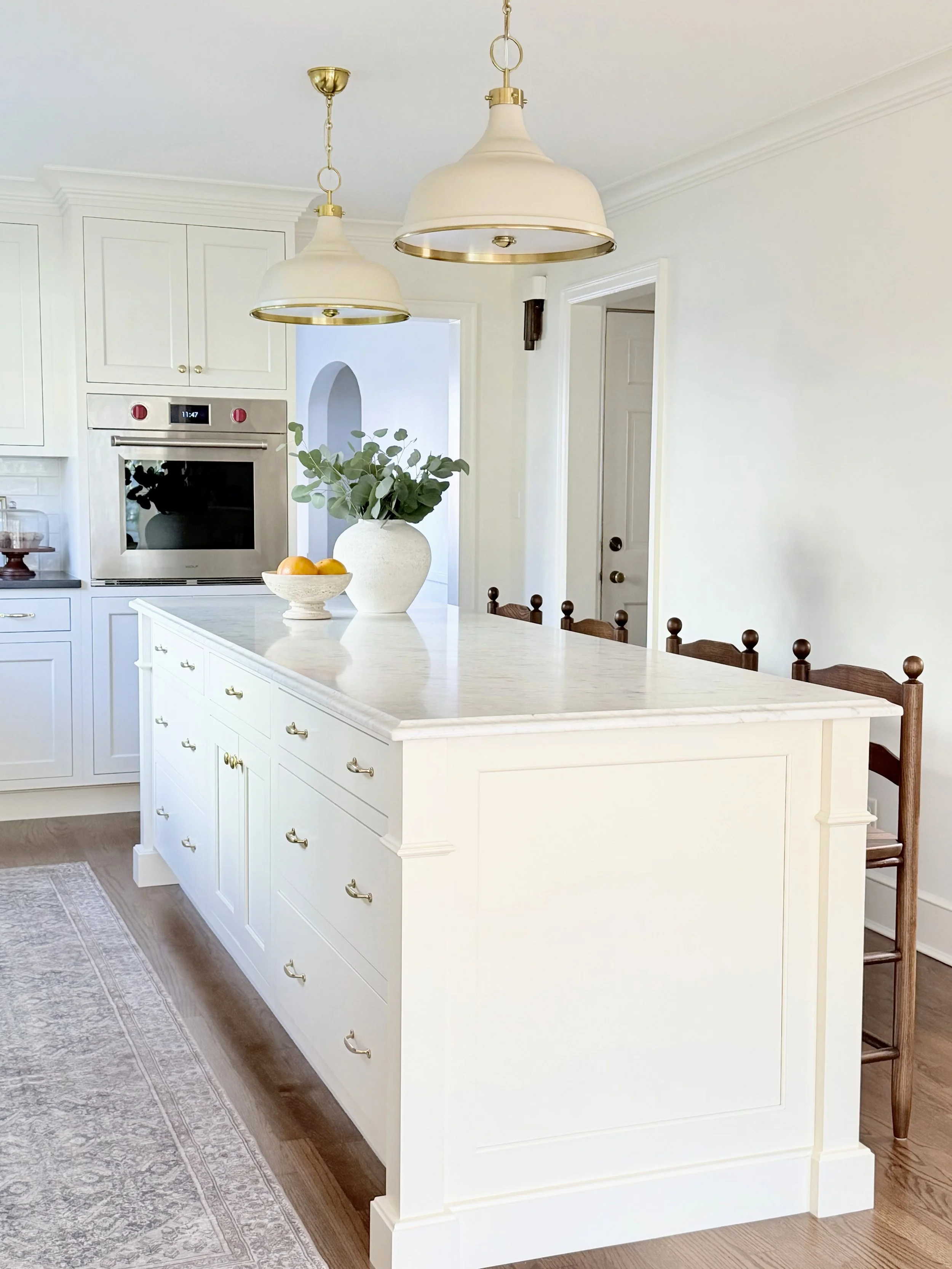 Bright white kitchen with island, beige pendant lights, vase with greenery, bowl of oranges, oven, and wooden chairs at the island.