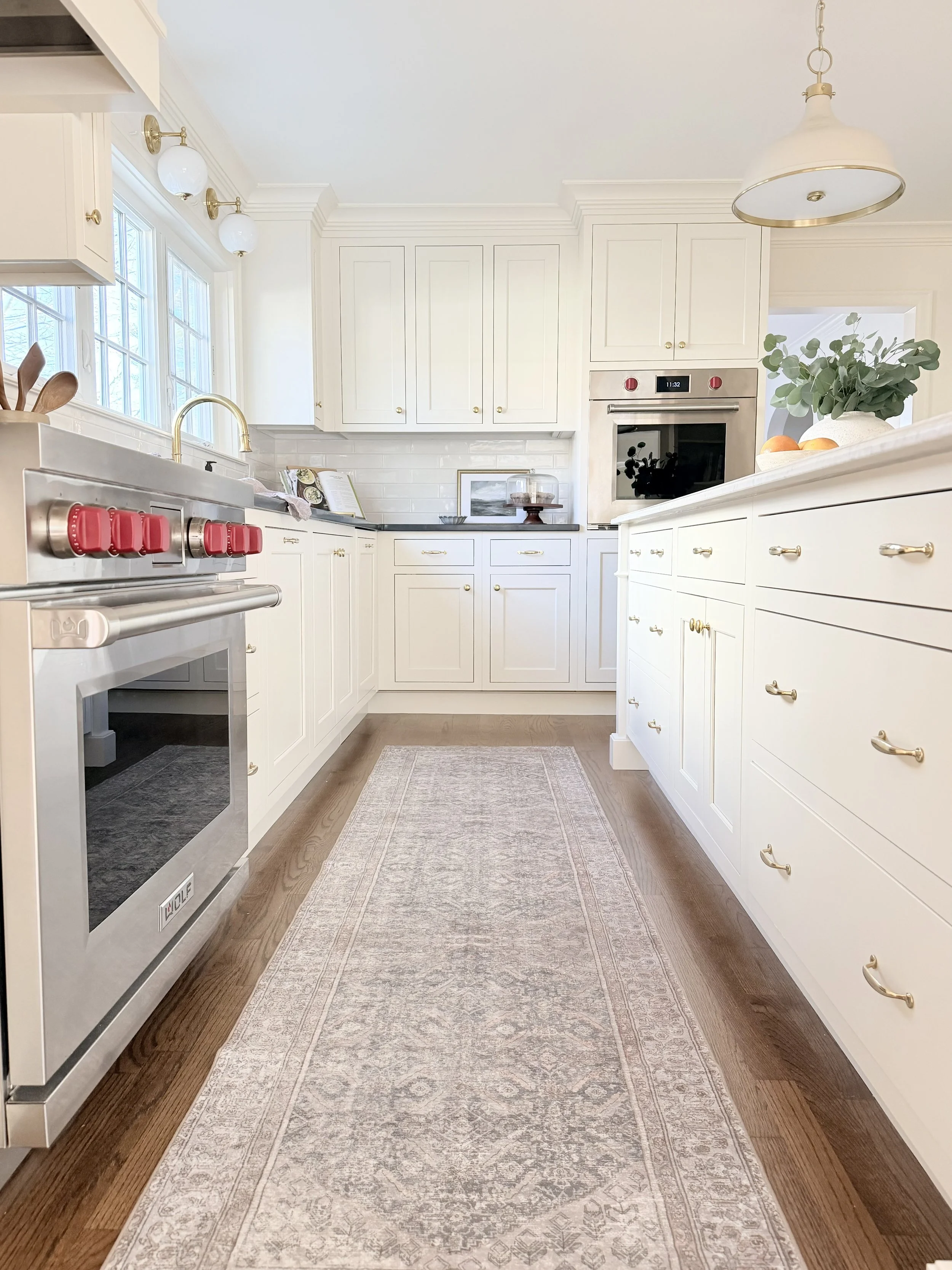 A bright white kitchen with white cabinetry, gold hardware, a stainless steel stove with red knobs, a built-in oven, a gold faucet, a window with multiple panes, a white and gold pendant light, a beige rug on wooden floor, and green foliage with peaches in a white bowl on the counter.