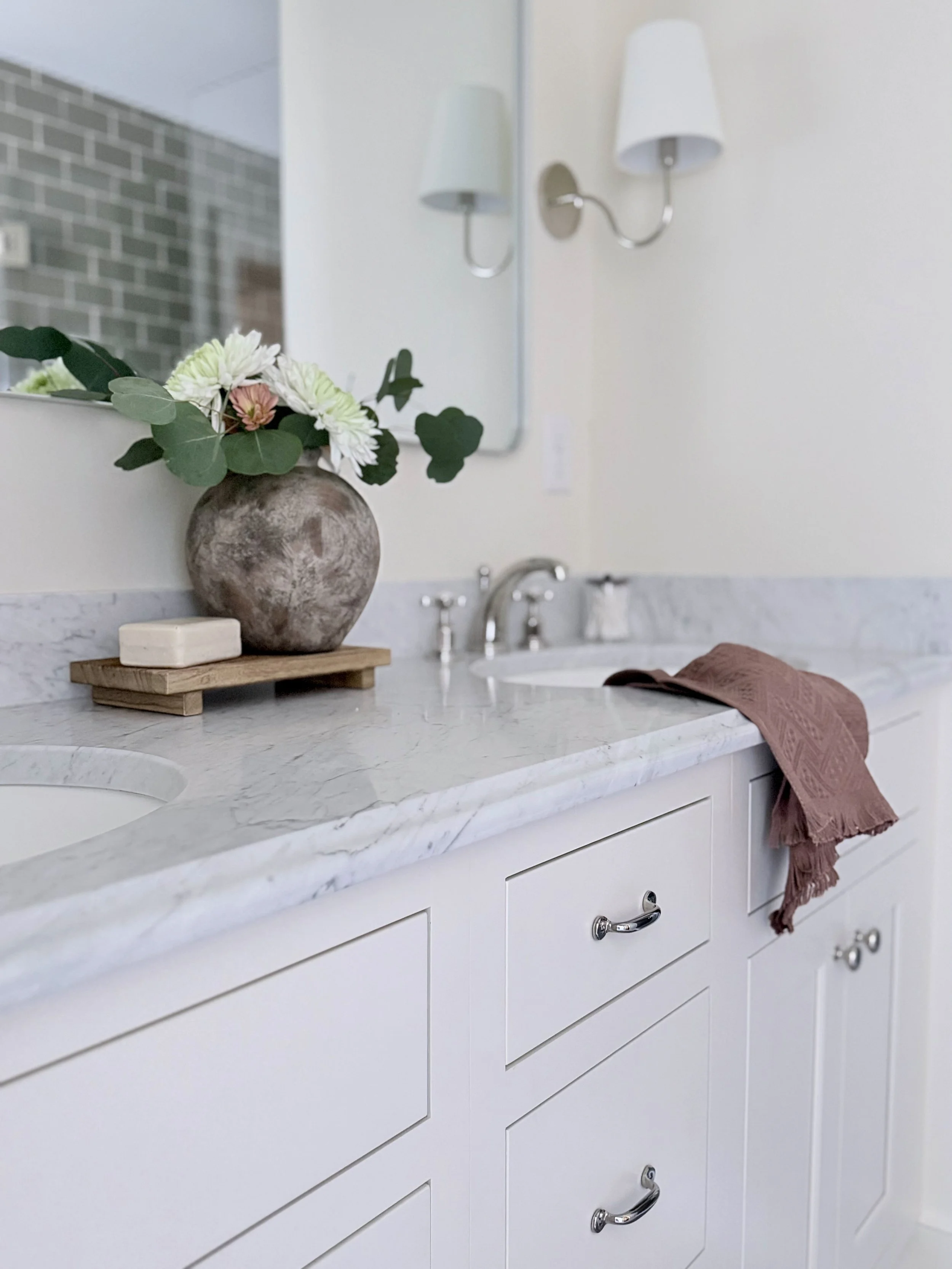 Bathroom with white vanity, marble countertop, a vase with flowers, a soap dish, a pink towel on the counter, and a mirror with wall-mounted lamps.