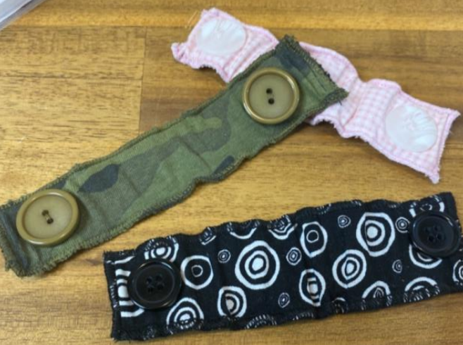 Two fabric wristband straps with large buttons, one in camouflage and pink pattern, the other in black with white circular patterns, placed on a wooden surface.